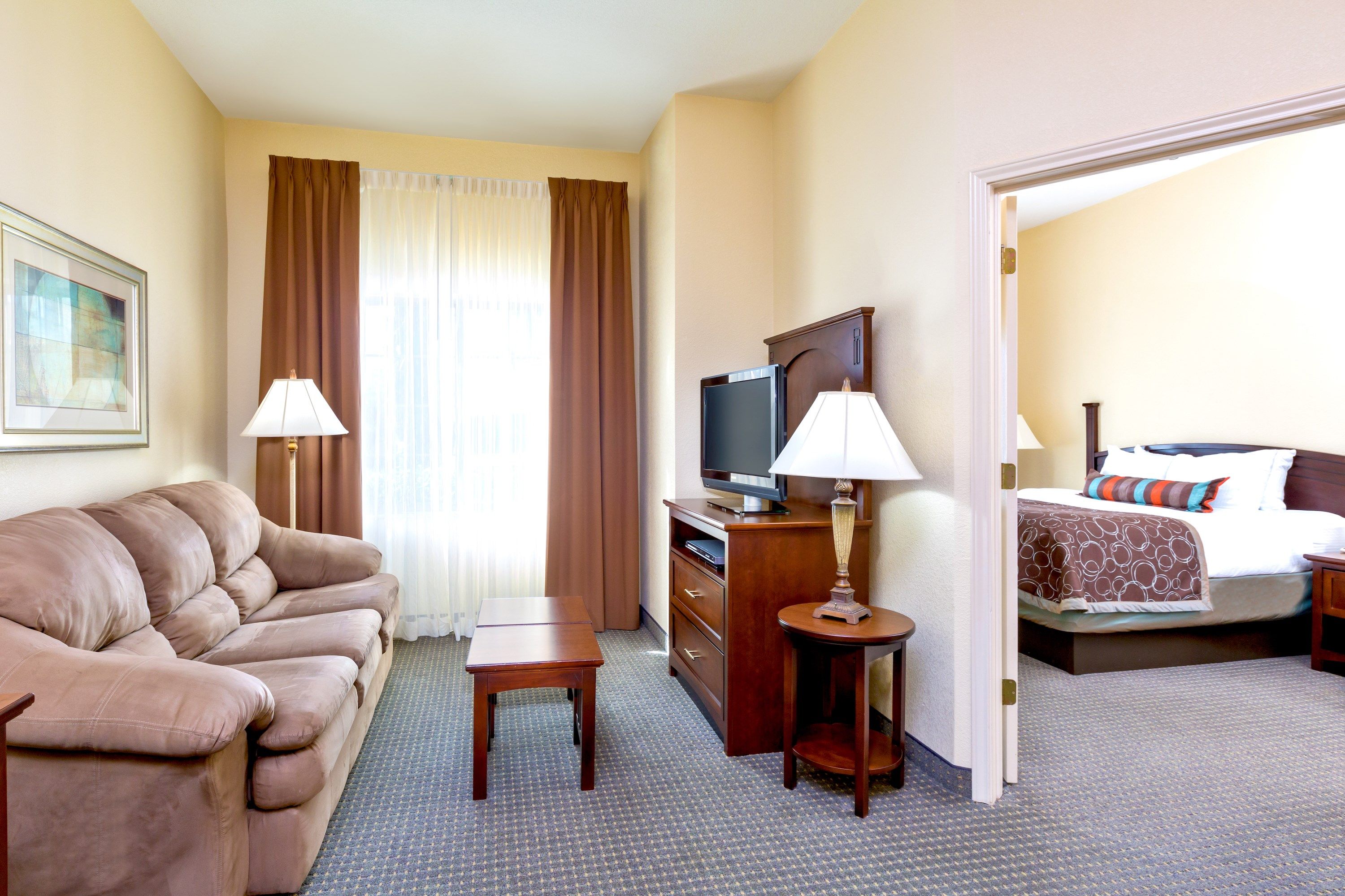 image Living area with beige sofa, coffee table, TV on wooden cabinet, and a lamp — doorway leads to the bedroom beyond