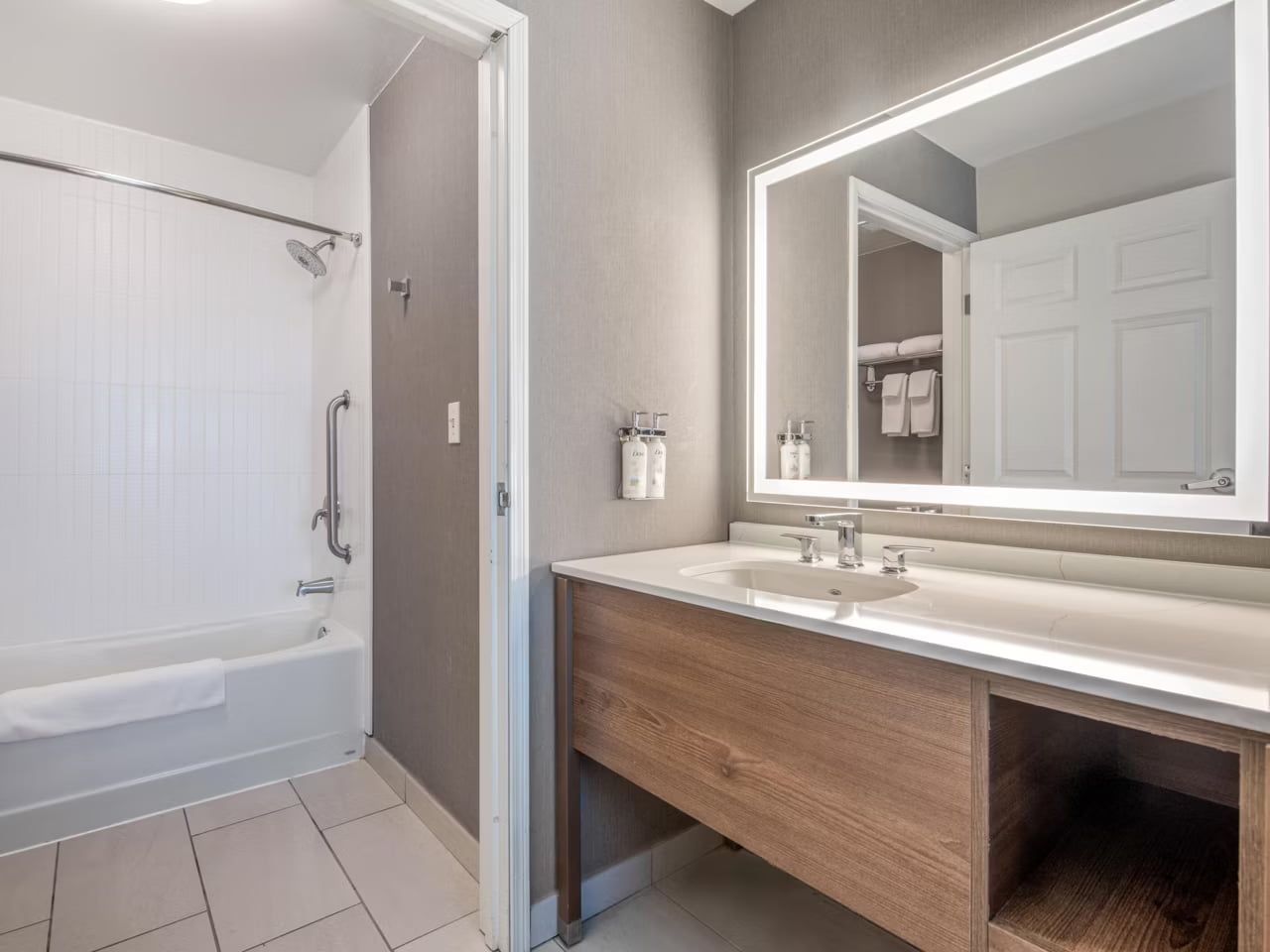 image A bathroom with a tub/shower combination, a grab bar, and a wood-tone vanity beneath a backlit mirror.