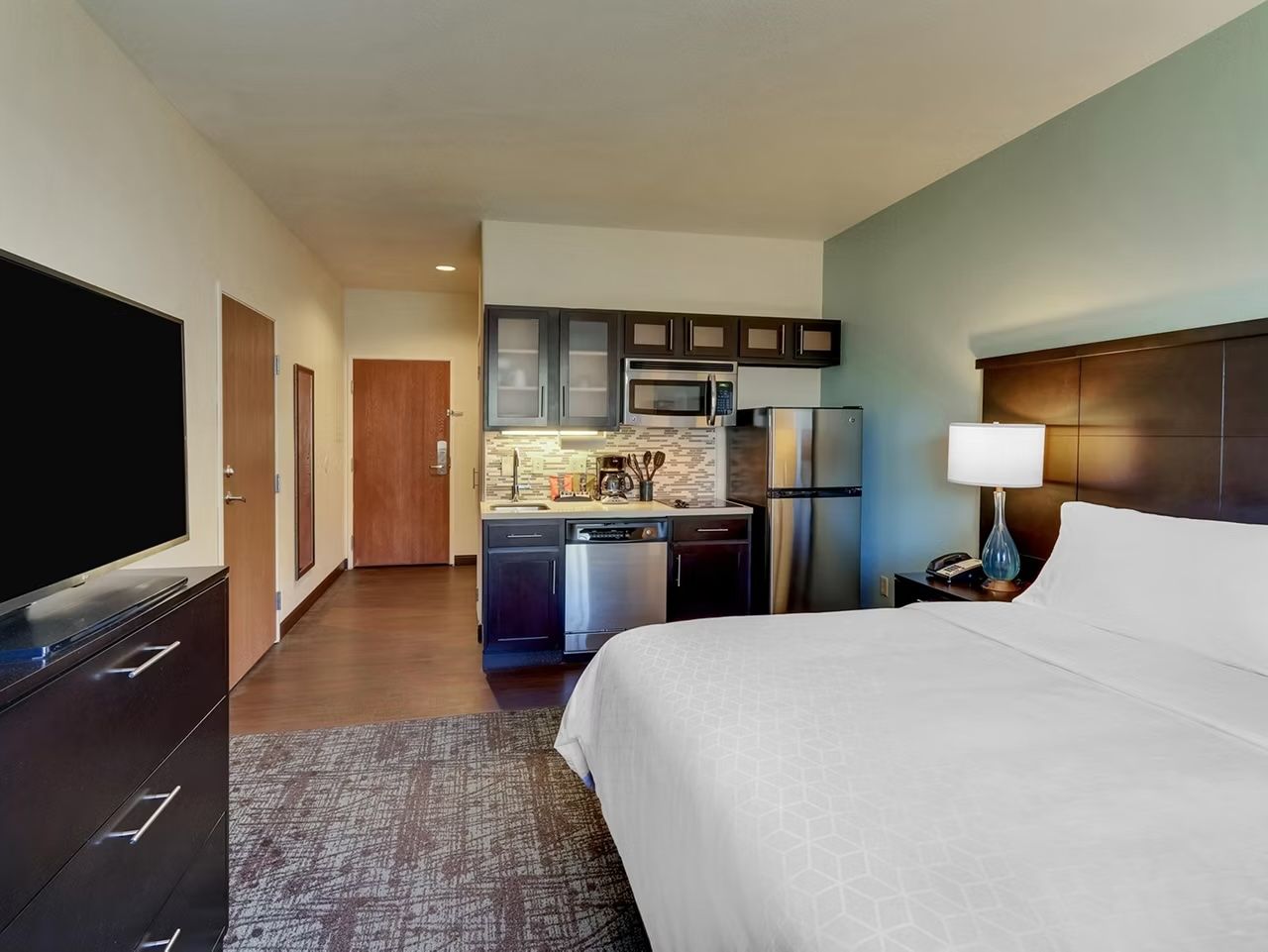 image Open-plan studio with king bed, kitchen with refrigerator and microwave, wall-mounted flat-screen TV, and dresser.