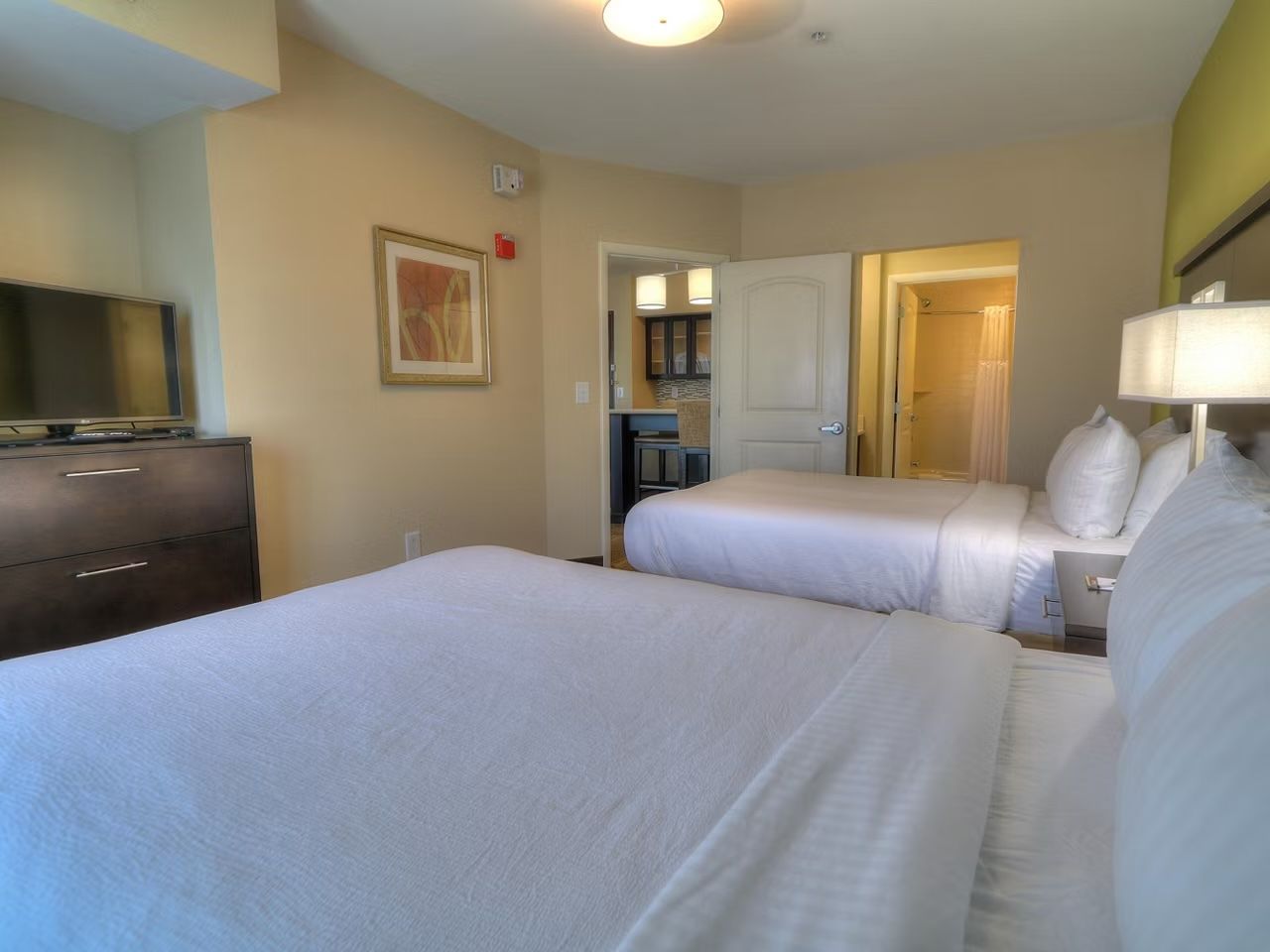 image Two beds with white linens and dark wood dresser — kitchenette and bathroom just through the doorway