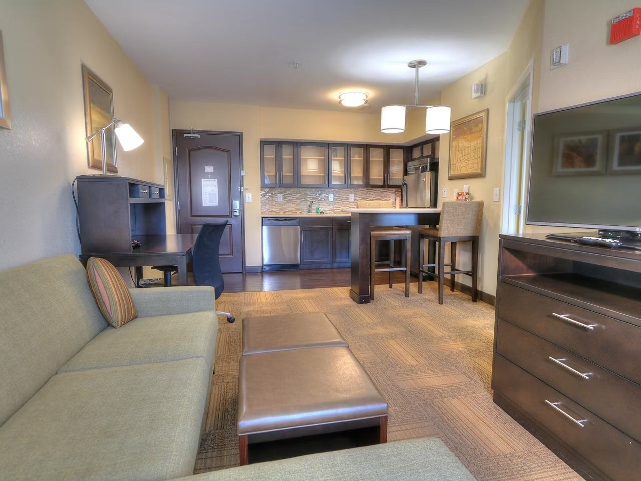 image Second living area angle — sofa, ottoman, and flat-screen TV with the kitchen just behind