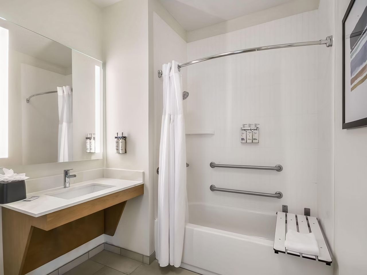 image Accessible bathroom with a grab-bar bathtub, wall-mounted toiletry dispensers, floating wood vanity, and an illuminated mirror