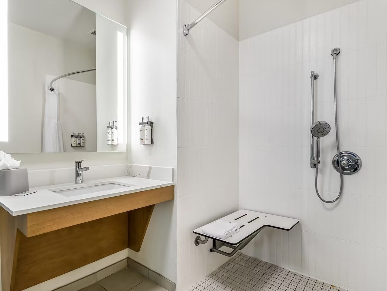 image Accessible bathroom with a roll-in shower, grab bars, built-in seat, floating wood vanity, and an illuminated mirror