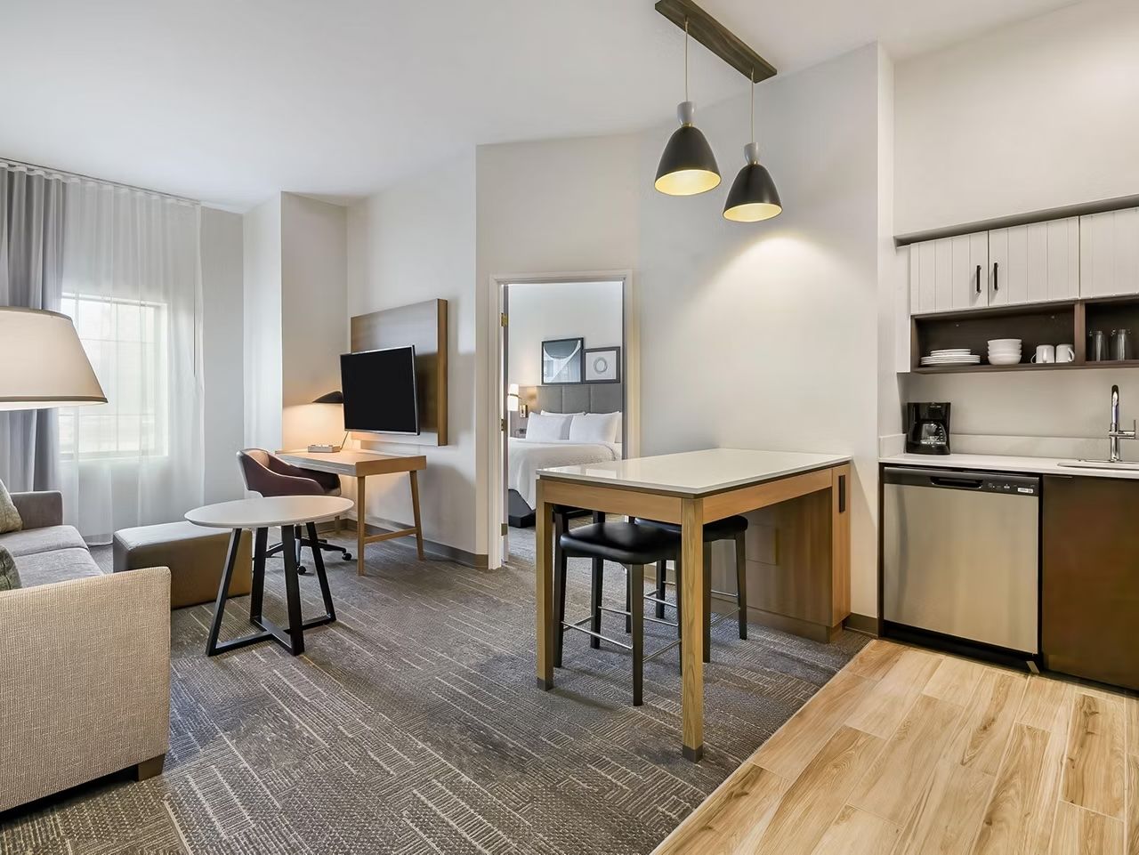 image Open-plan living area with a sofa, coffee table, dining table, kitchen, and wall-mounted TV — bright with natural light