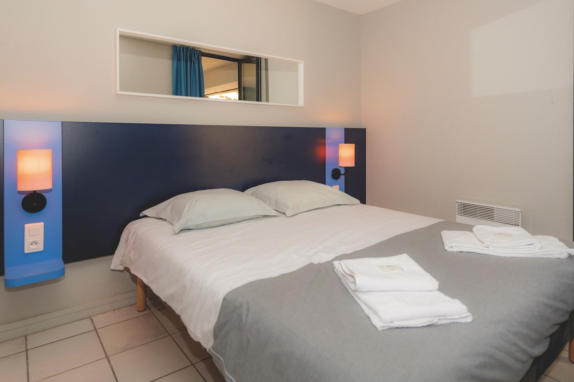 image A dark blue headboard, a wall lamp on each side and a mirror above frame the bed with grey and white bedding