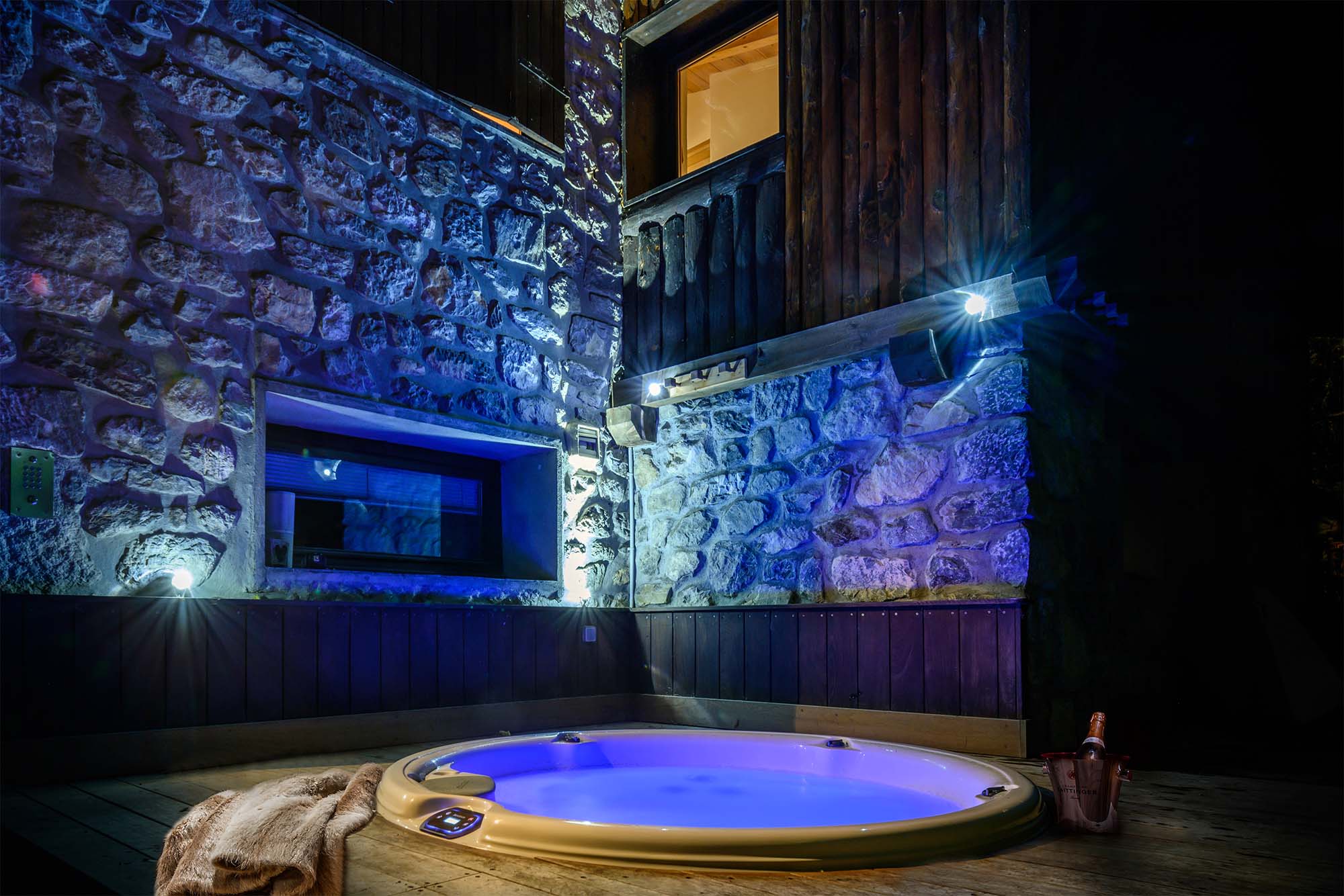 image Unwind in the outdoor hot tub, set against a beautiful stone wall and illuminated by soft blue lighting. This private retreat is perfect for soaking under the stars and enjoying the tranquil sounds of nature.