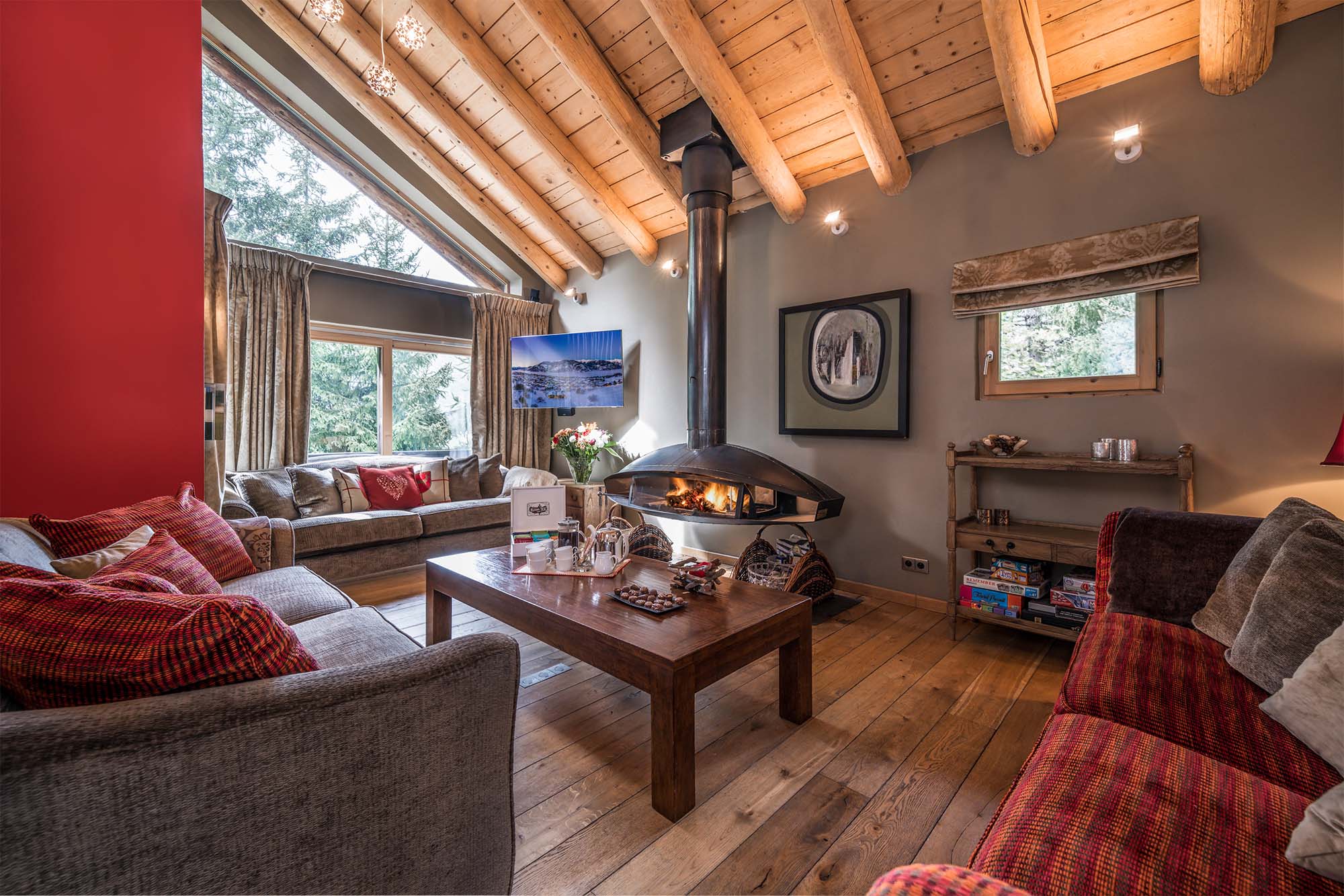 image Relax in this cozy living room featuring plush seating, vibrant red accents, and a rustic wooden ceiling. Large windows flood the space with natural light, creating a warm and inviting atmosphere perfect for unwinding after a day of exploring.