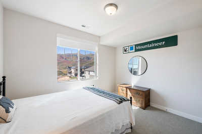 A sunlit queen bedroom with a panoramic window offering a scenic mountain view, complemented by a classic wooden chest and a neatly draped bed.