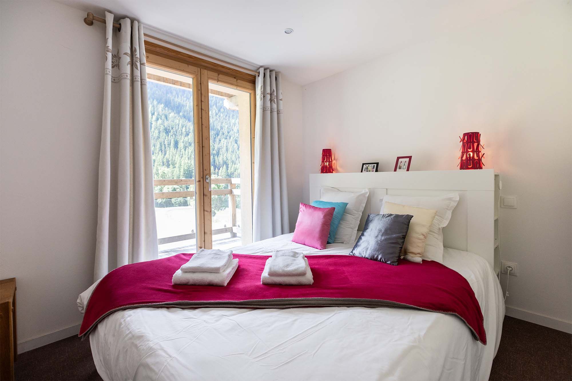 image Rest peacefully in this cozy bedroom featuring a comfortable double bed with soft linens and vibrant red accents. Large windows offer plenty of natural light and serene views of the outdoors.