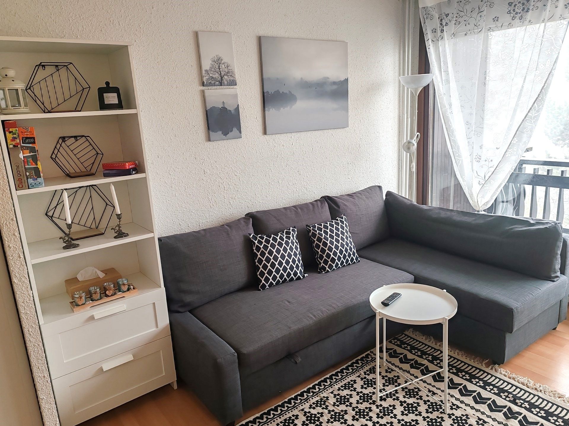 image Relax in this stylish living room featuring a comfortable sectional sofa that easily converts into a sofa bed, a chic bookshelf with decorative accents, and large windows that flood the space with natural light.