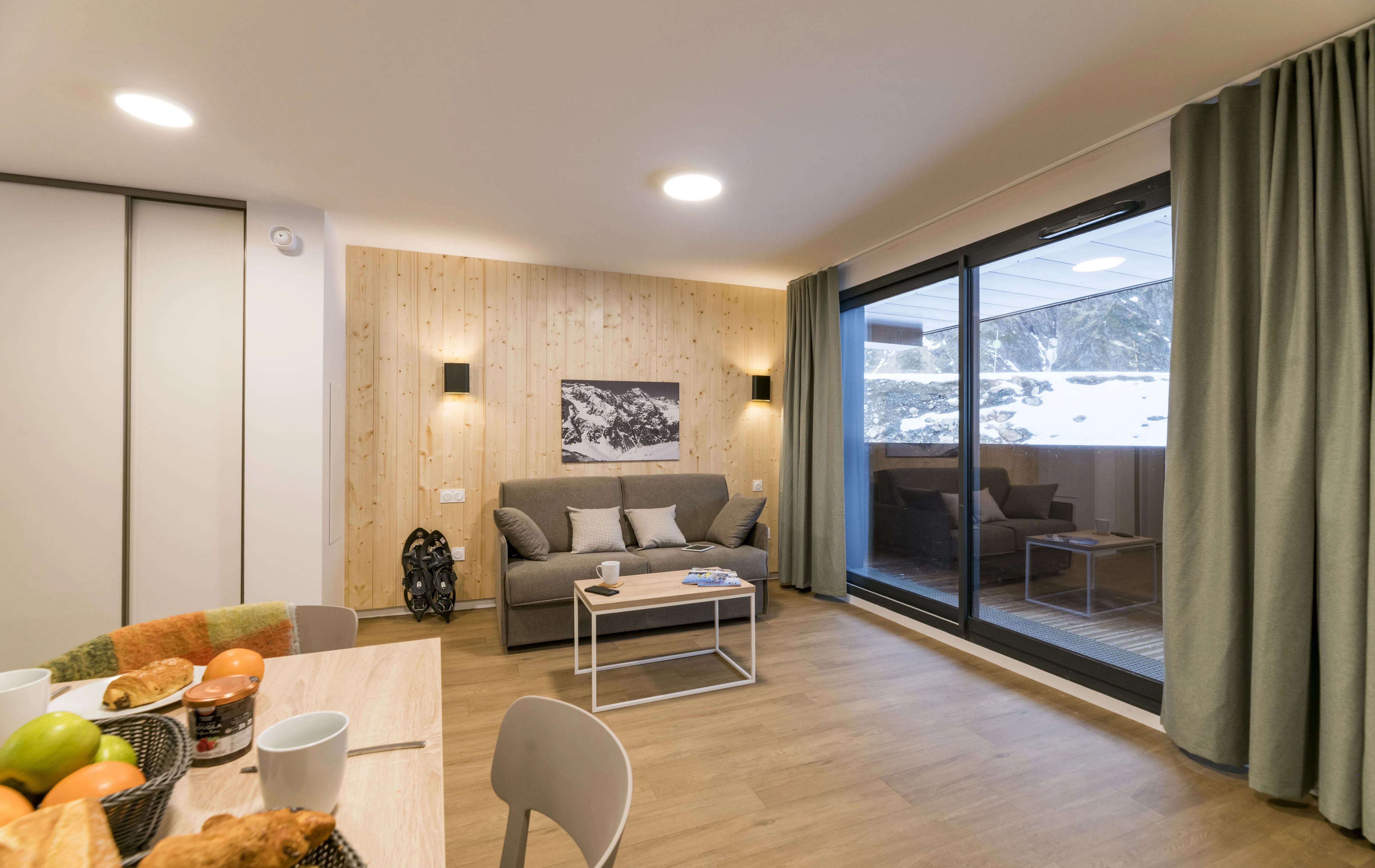 image Relax in this cozy living space with wooden accents, perfect for family gatherings after a day on the slopes, featuring large windows that offer a view of the snow-covered outdoors.