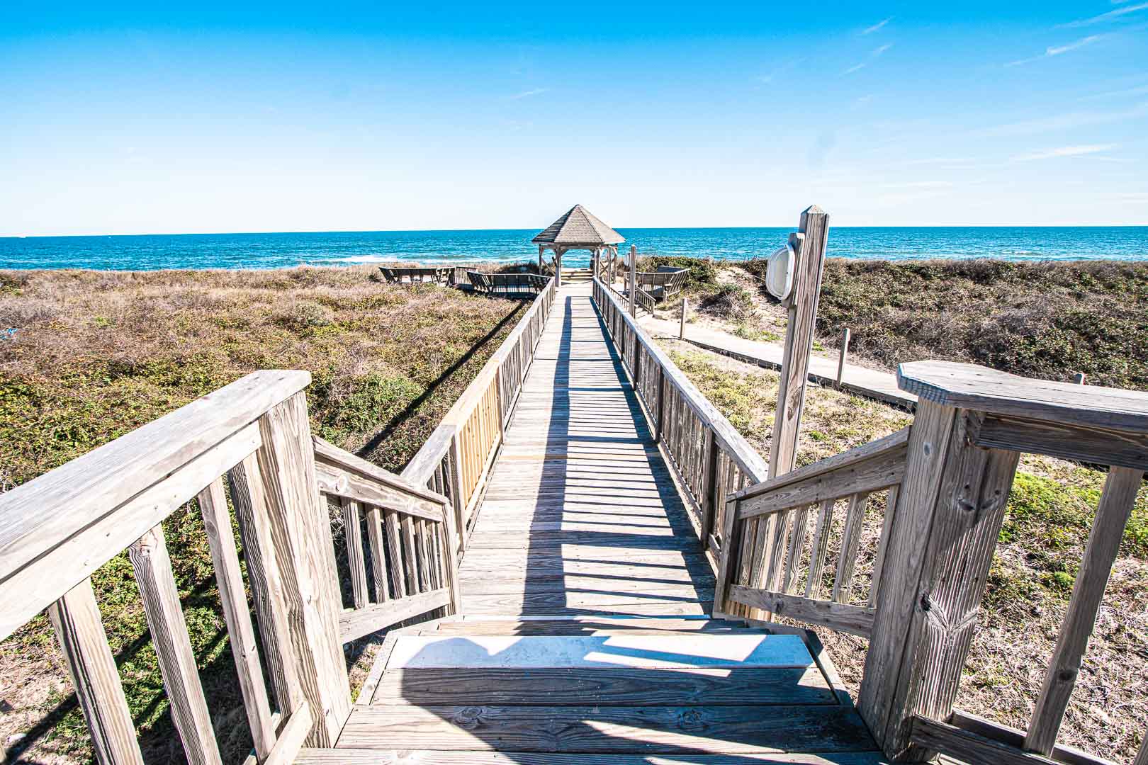 image A serene wooden boardwalk leading to the beach, with a gazebo at the end and a peaceful coastal landscape in the background.