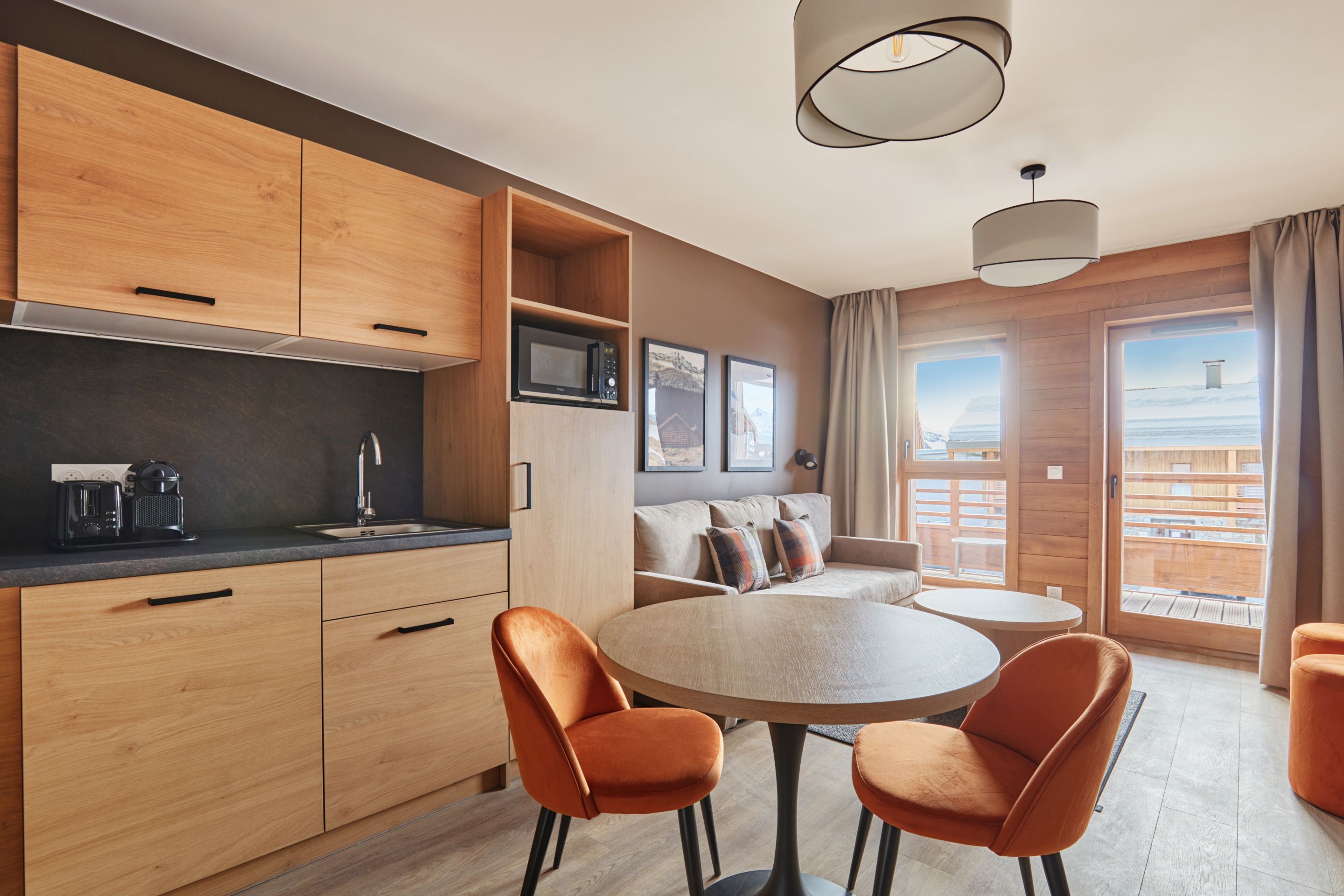 image Open-plan living and dining area with orange velvet chairs, a round table, grey sofa, and a kitchenette with light wood cabinetry.