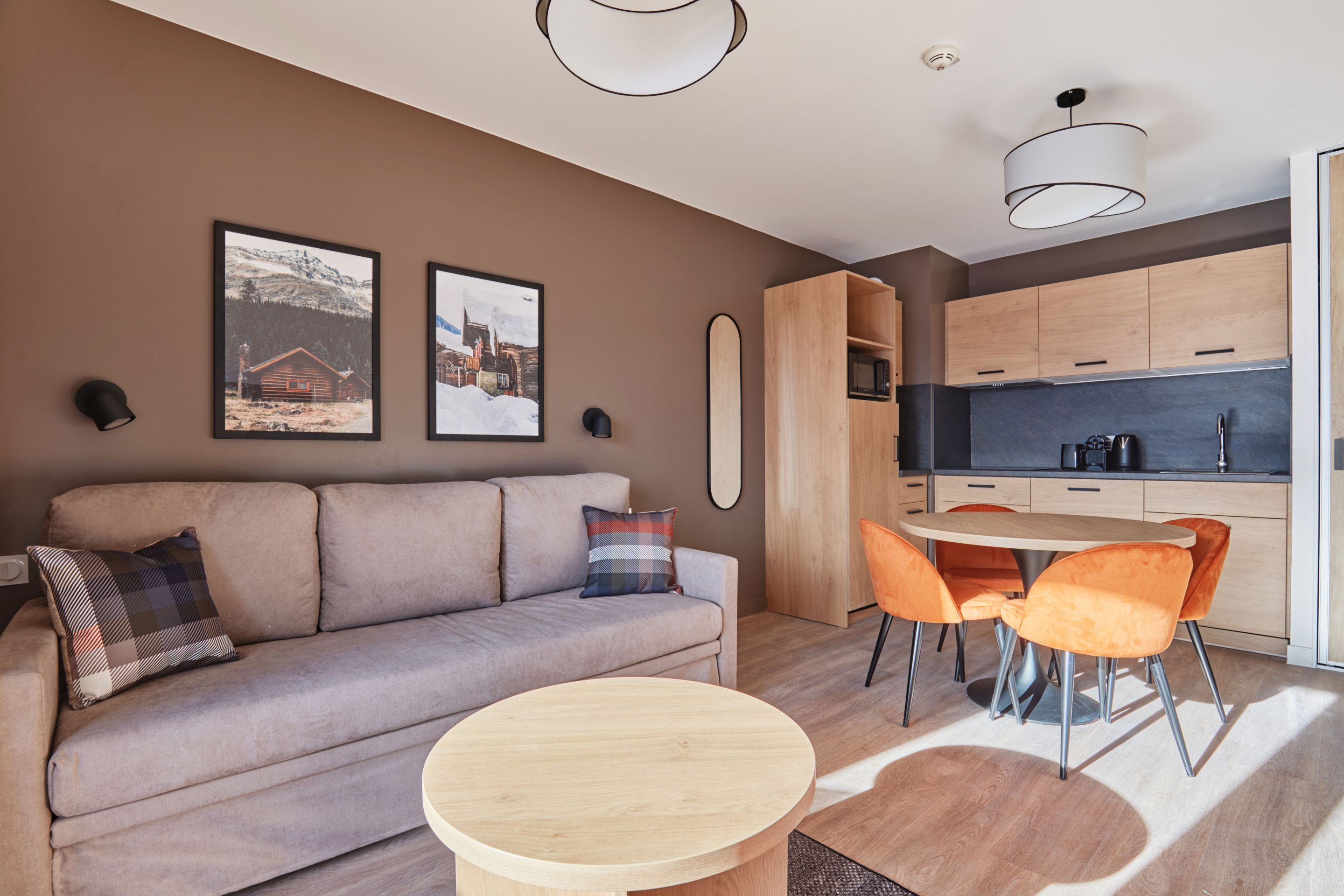 image Living and dining area with a grey sofa, round table, orange chairs, and a kitchenette with light wood cabinetry and dark countertop.