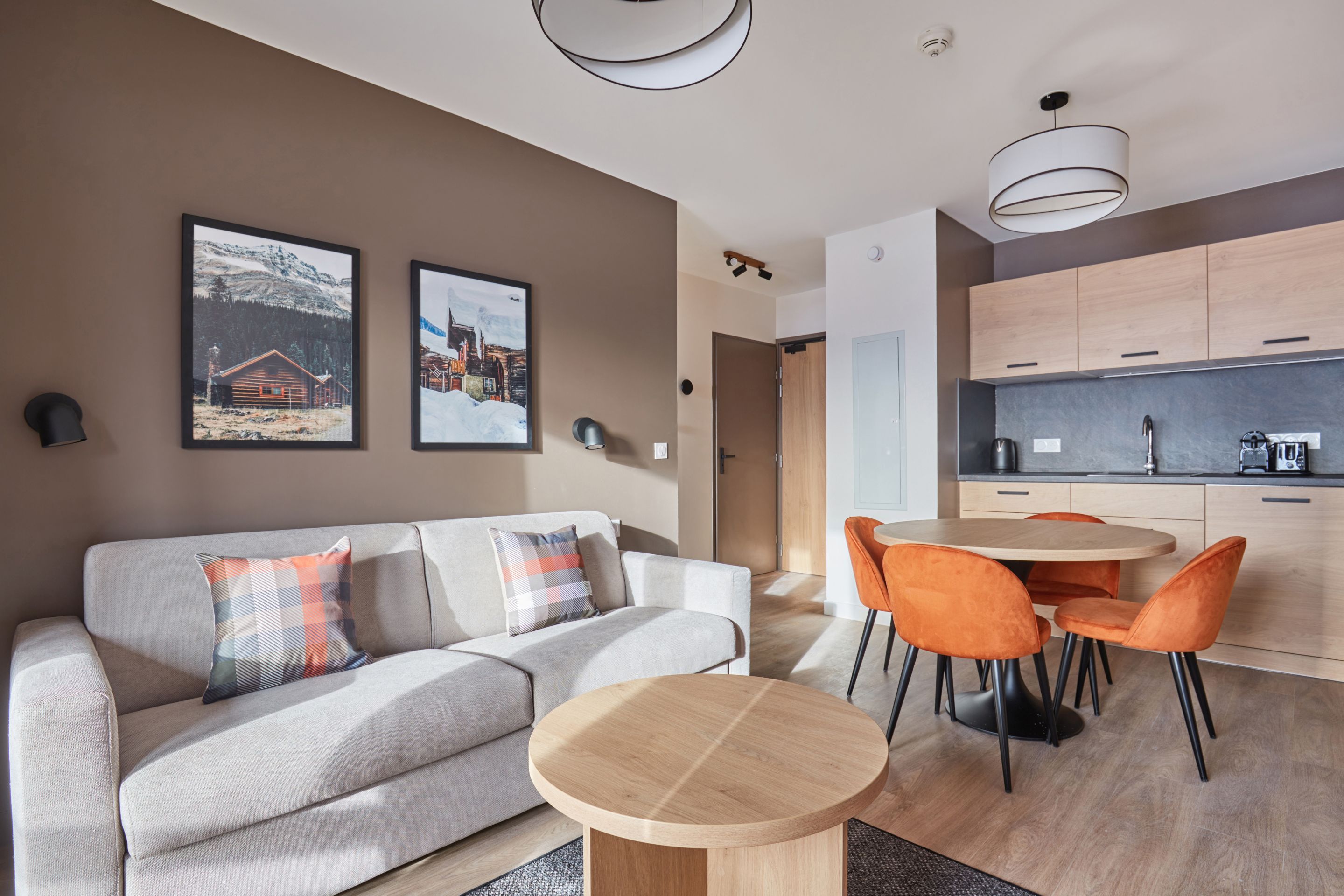 image Open-plan living and dining area with grey sofa, round table, orange chairs, and a kitchenette with light wood cabinetry.