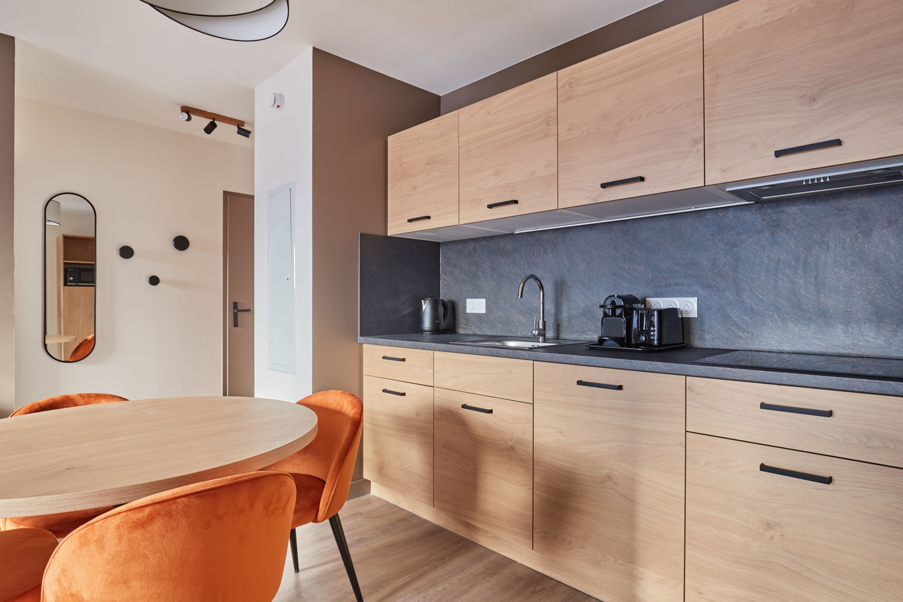 image Kitchenette close-up with light wood cabinetry, dark stone countertop, sink, and orange dining chairs beside the round table.