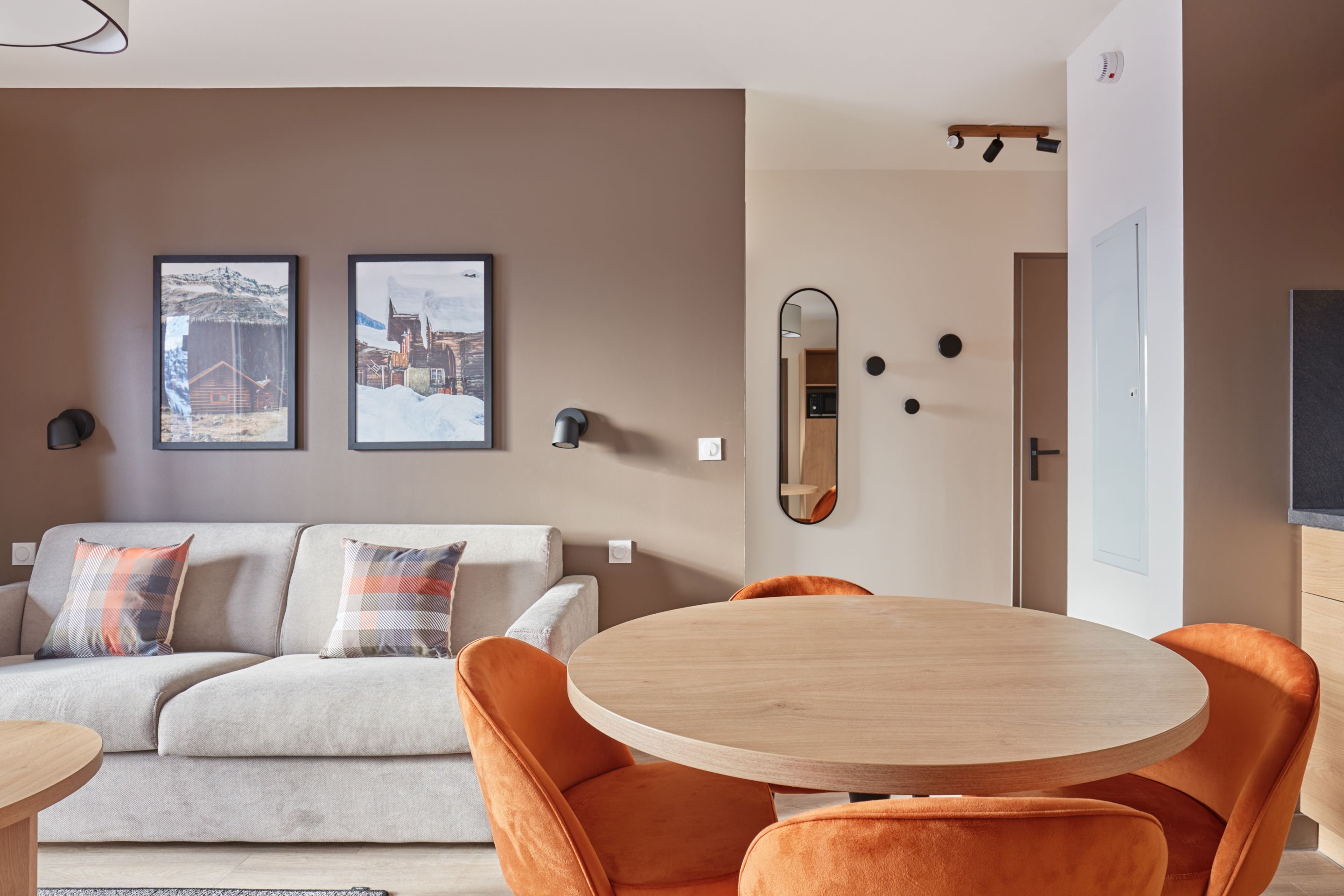 image Dining area with round table and orange chairs alongside framed landscape prints on a warm brown wall.