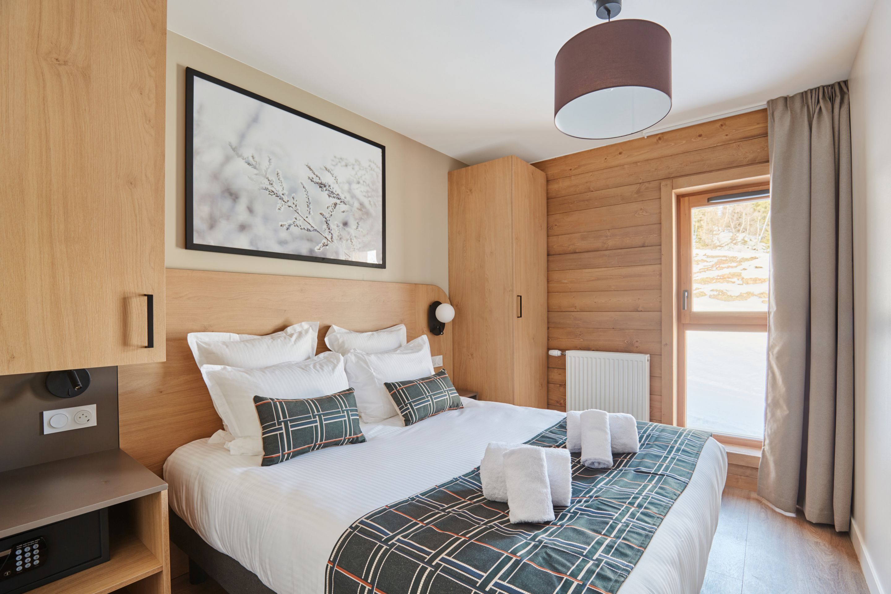 image Double bedroom with an upholstered headboard, white linens, plaid pillows, wooden wardrobe, and a window with natural light.