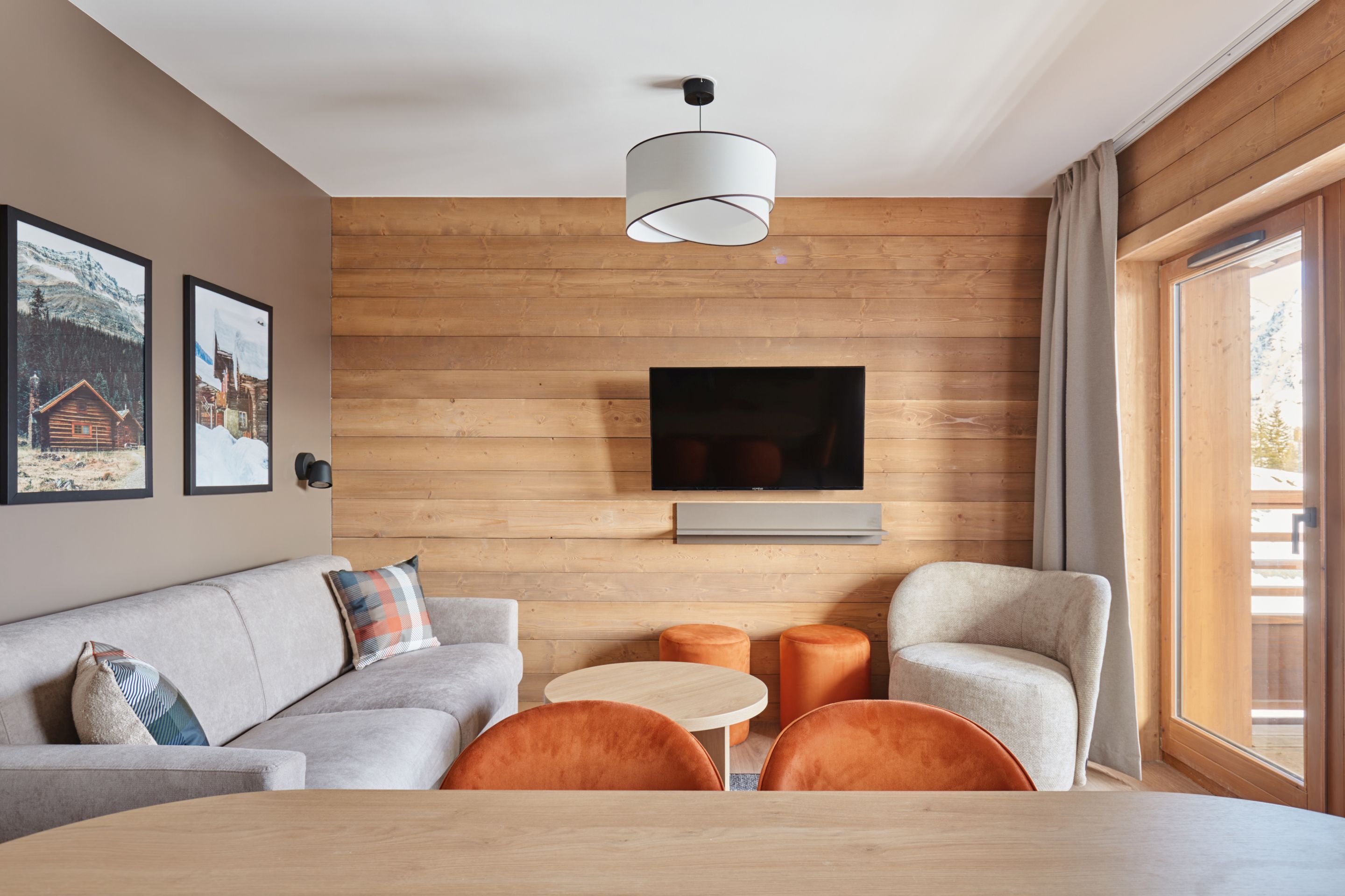 image Living area with a wood-panelled wall, grey sofa, mounted TV, and a large balcony door with beige curtains.