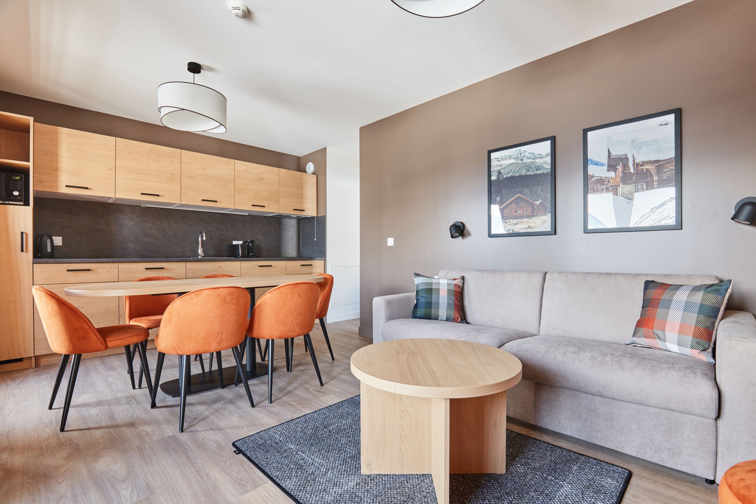 image Living and dining area with round table, orange chairs, grey sofa, and a kitchenette with light wood cabinetry and dark countertop.