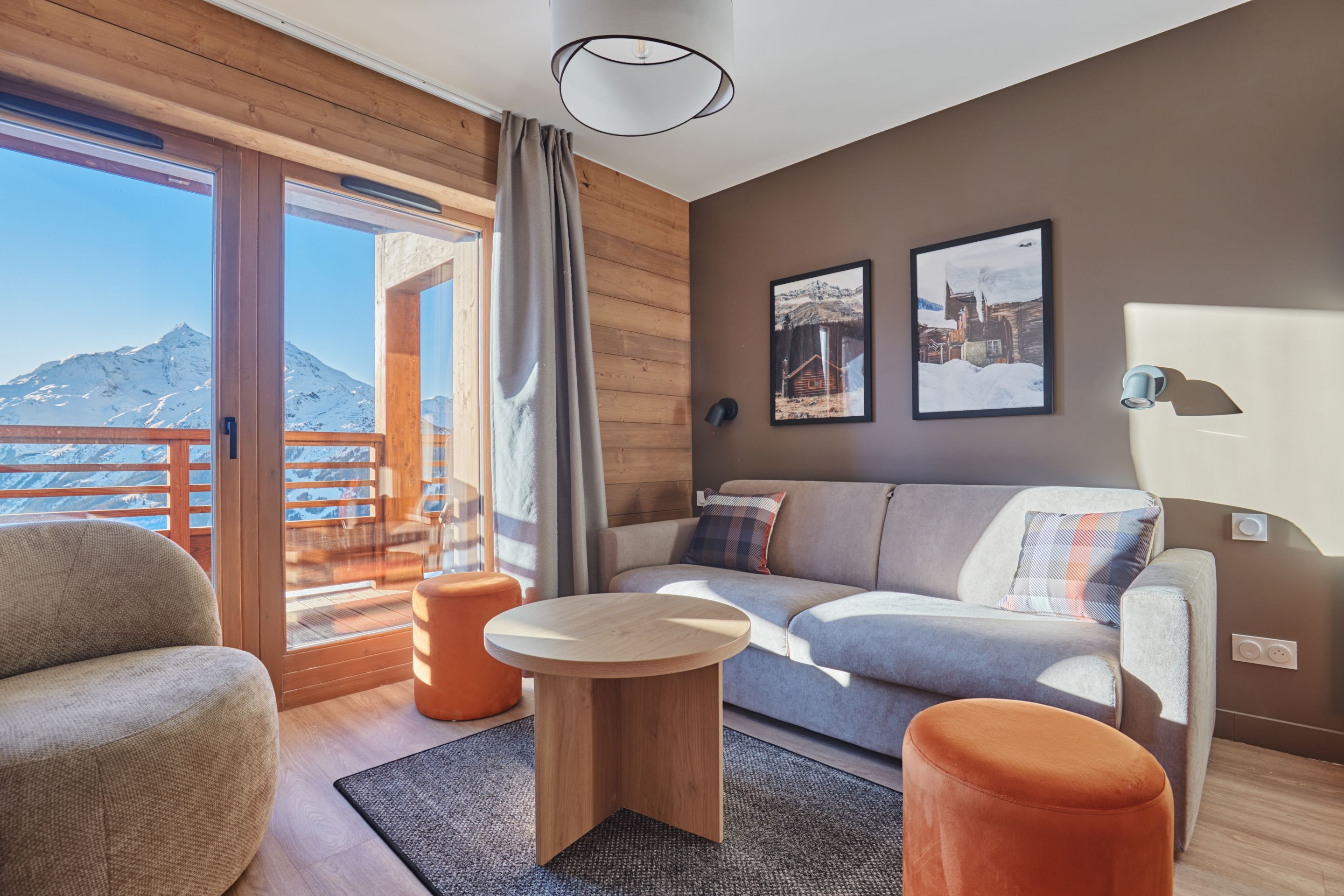 image Living area with a grey sofa, plaid cushions, orange stools, wooden wall panelling, and a large window framing mountain views.