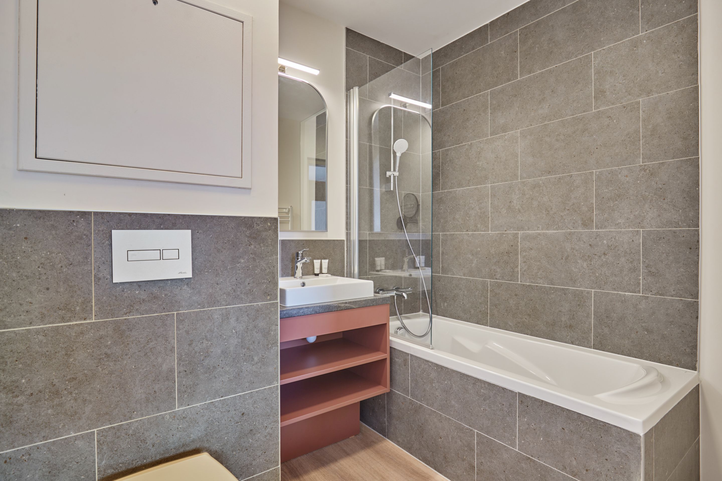 image Grey-tiled bathroom with a bathtub, glass shower screen, and modern sink — towels included.