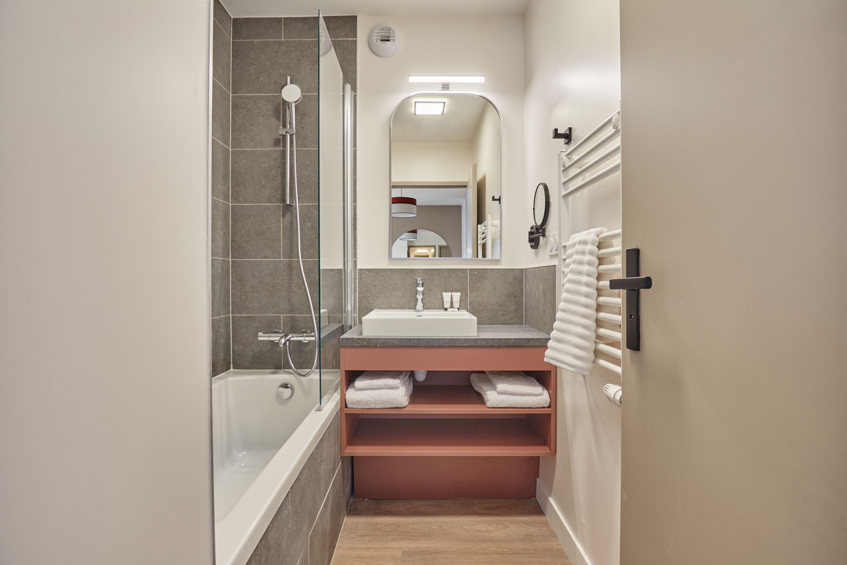 image Grey-tiled bathroom with a bathtub, glass shower, and wooden shelving stocked with folded towels.