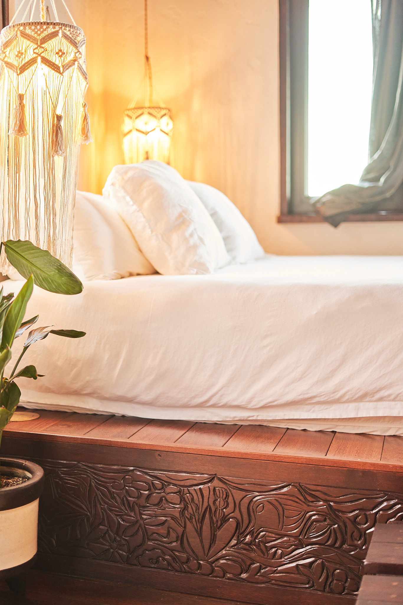 image Detailed wood-carved bed frame beneath glowing pendant lights and bright window views.