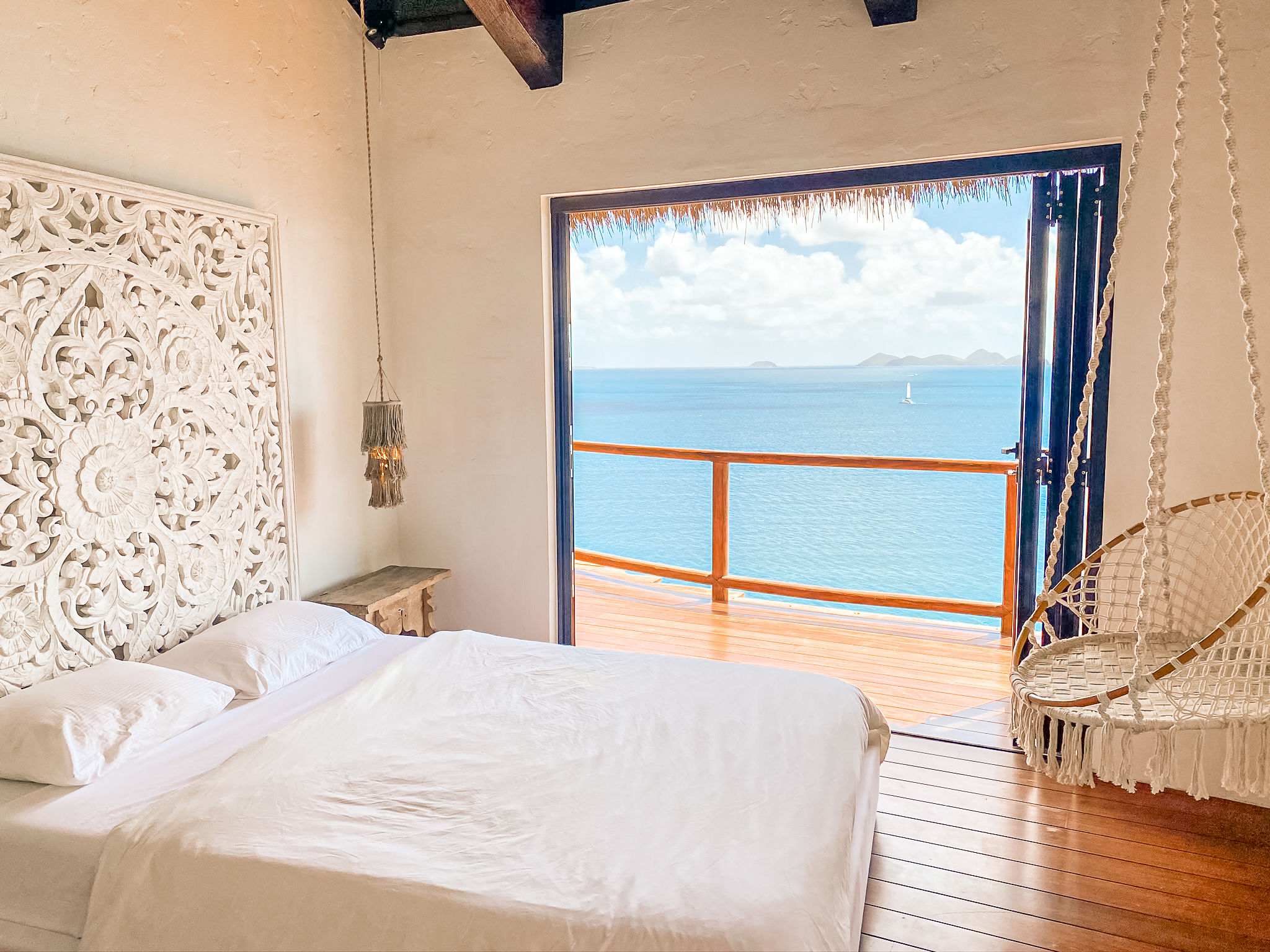 image Sunlit bedroom with ocean views framed by a decorative carved headboard and swinging chair.