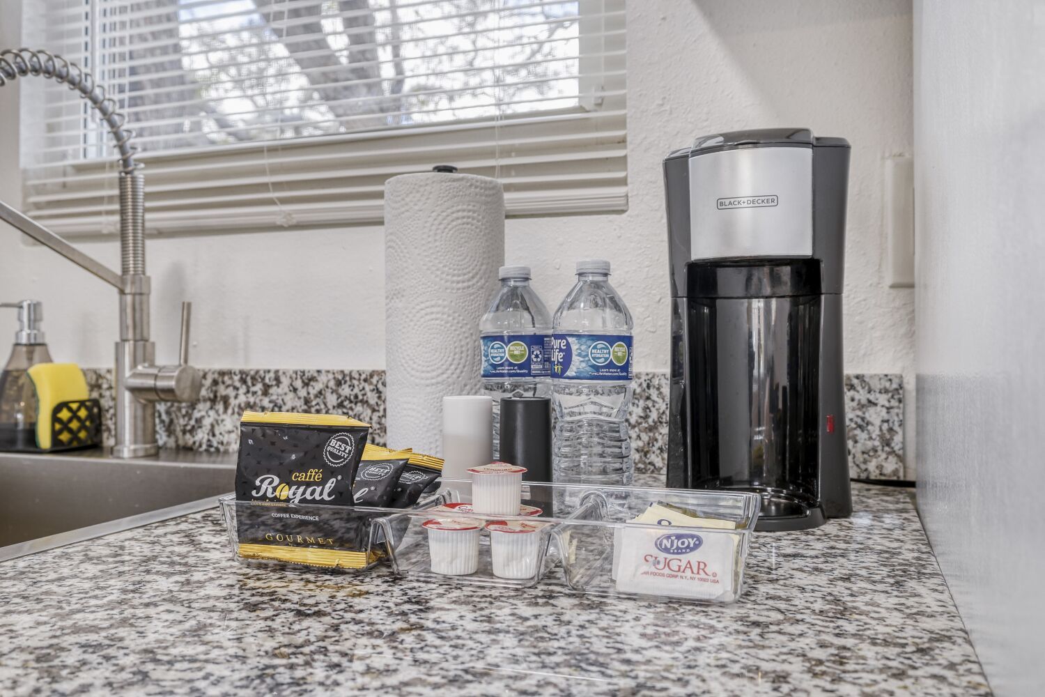 image Coffee setup on a granite kitchen counter with a coffee maker, bottled water, paper towels, and coffee pods beside the sink.