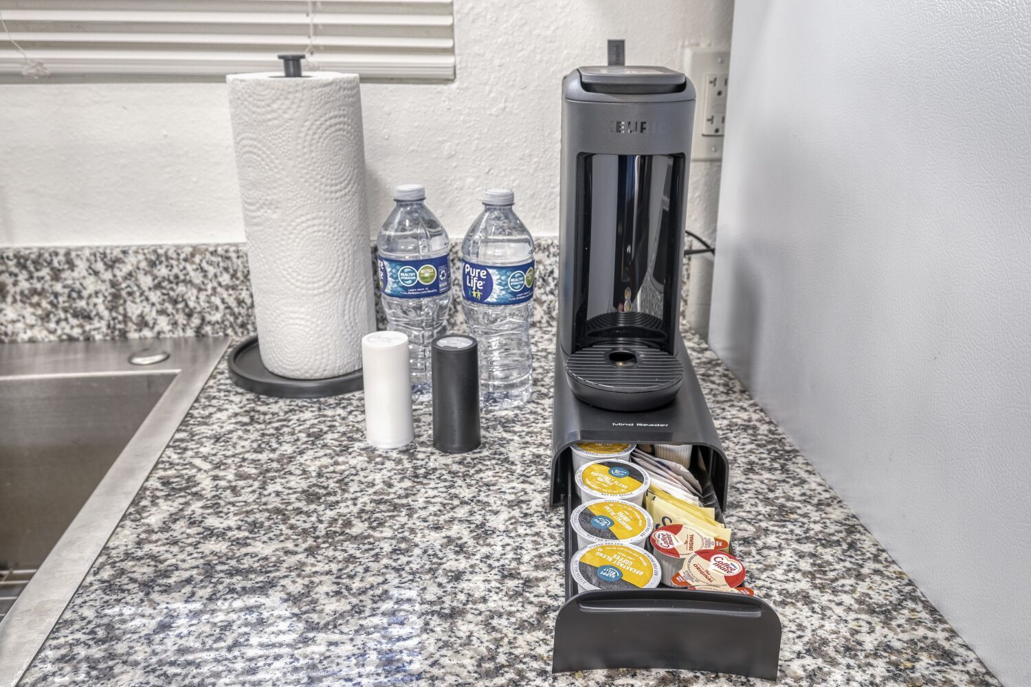 image Coffee station with a pod-style coffee maker and assorted coffee pods on a granite countertop next to bottled water and paper towels.