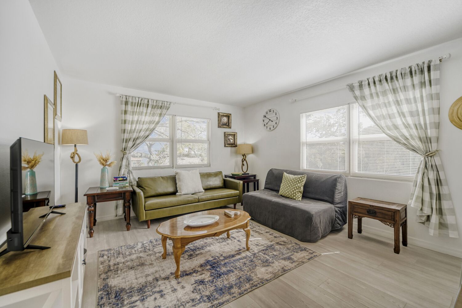 image A cozy living room features green and gray sofas, a wooden coffee table, and light curtains, with a decorative clock and wall art enhancing the inviting atmosphere.