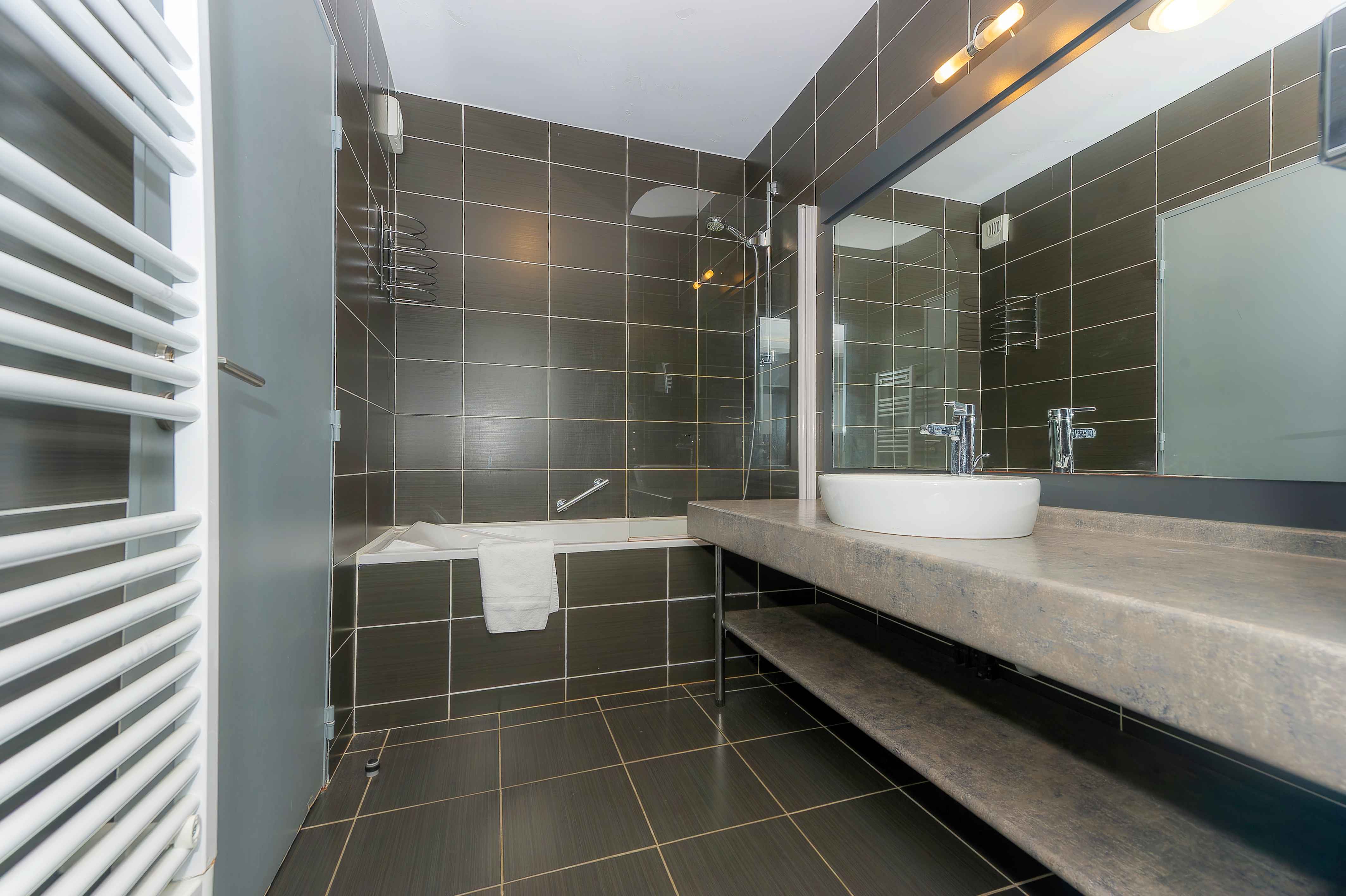 image Dark brown tiled bathroom with a bathtub and overhead shower, round vessel sink on a stone countertop, and a heated towel rail — fresh towels included.
