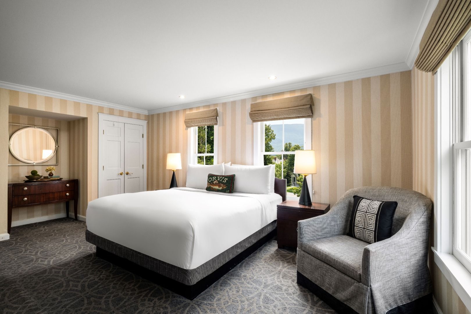 image A second inviting bedroom boasts a comfortable king bed with a decorative accent pillow, ambient lighting, and ample natural light streaming through the well-appointed windows and classic striped wallpaper.
