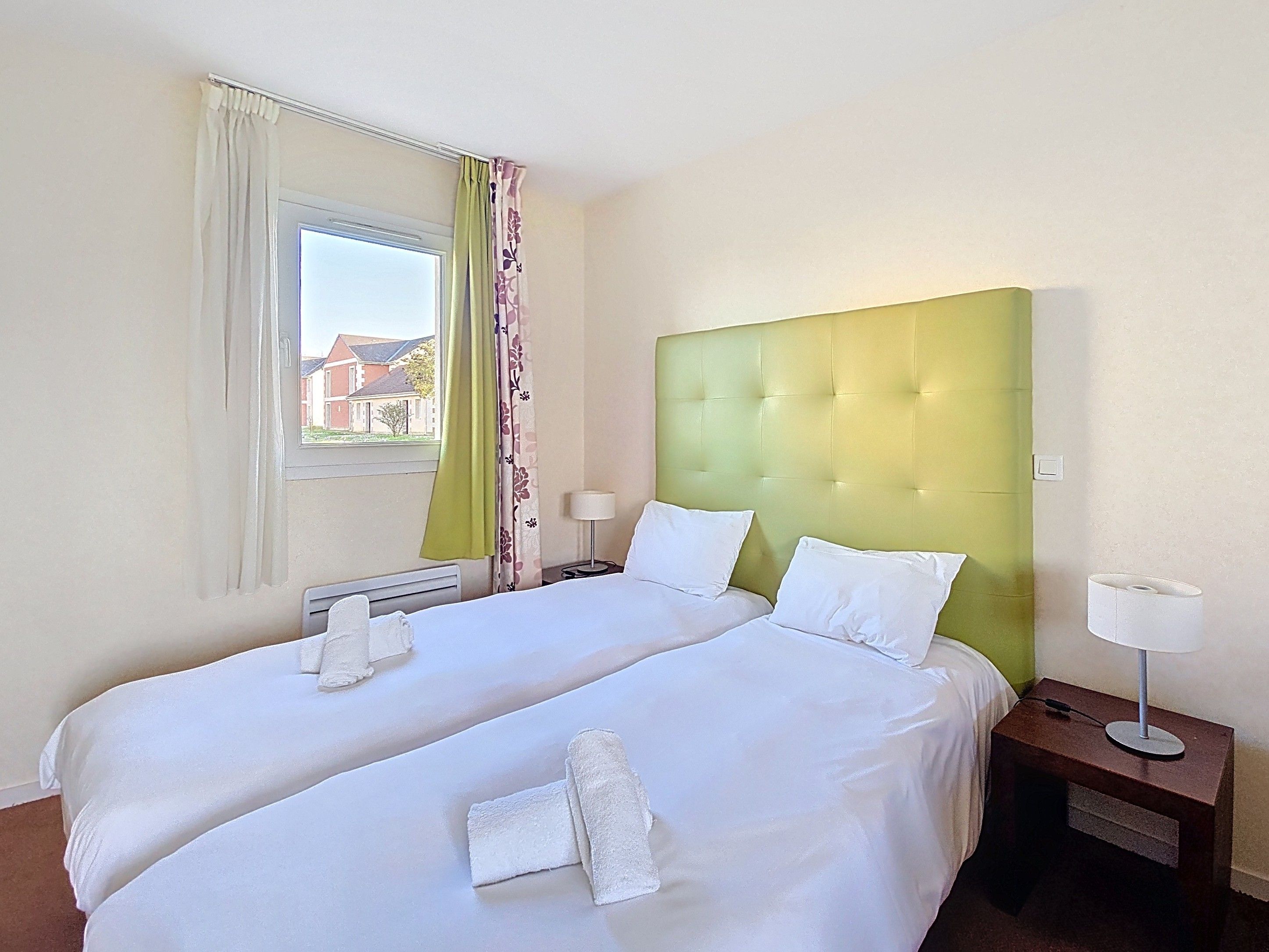 image Two single beds with a green padded headboard, white bedding, and a bedside lamp beside a window