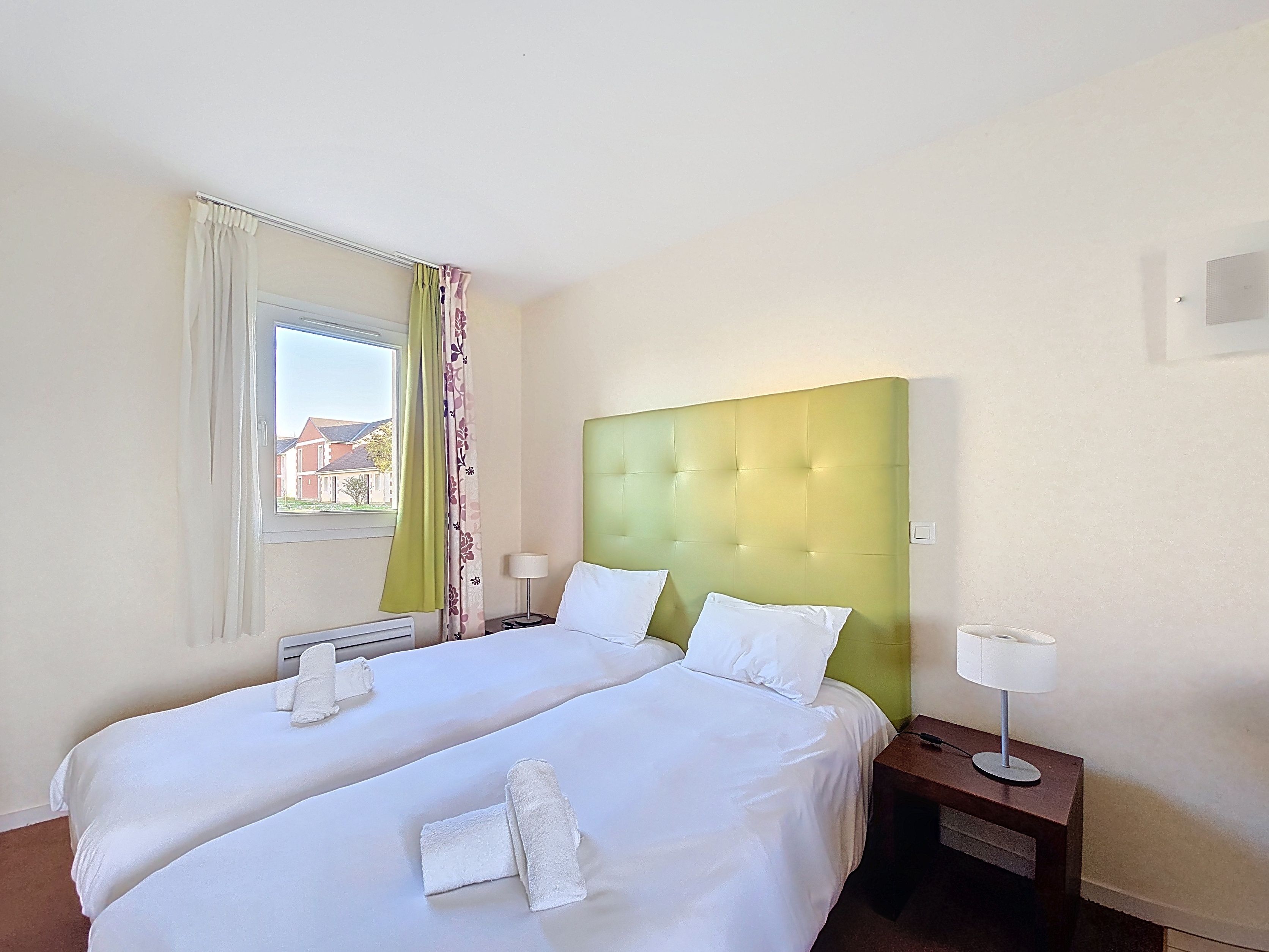 image Two single beds with green headboards and white bedding, each with a bedside lamp and natural daylight