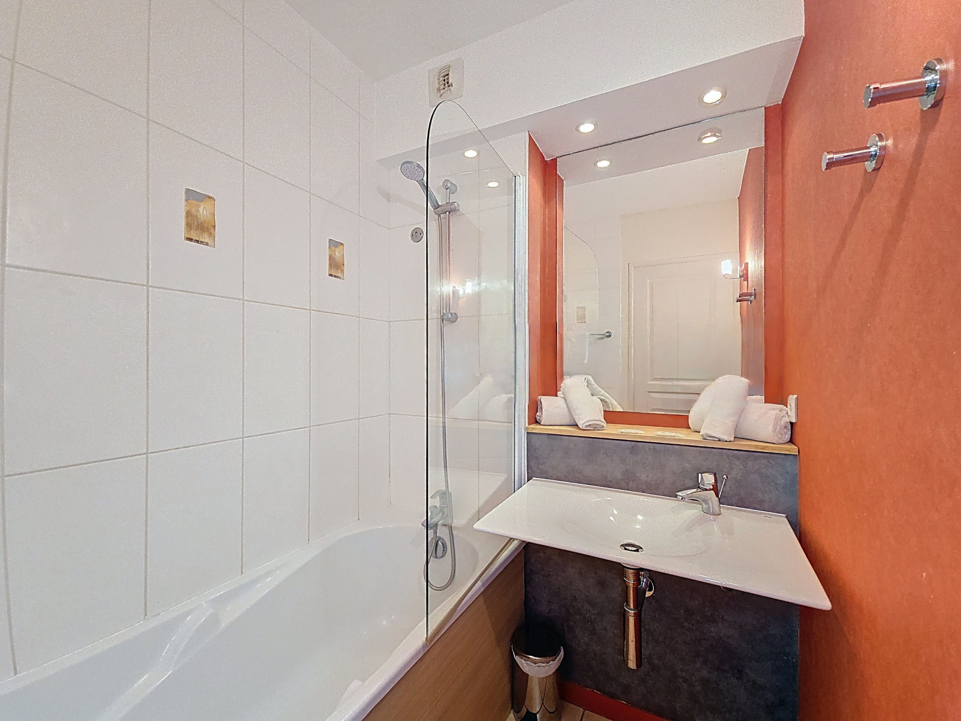 image Bathroom with white-tiled bathtub and shower attachment, modern tap, and towels laid out beside the tub