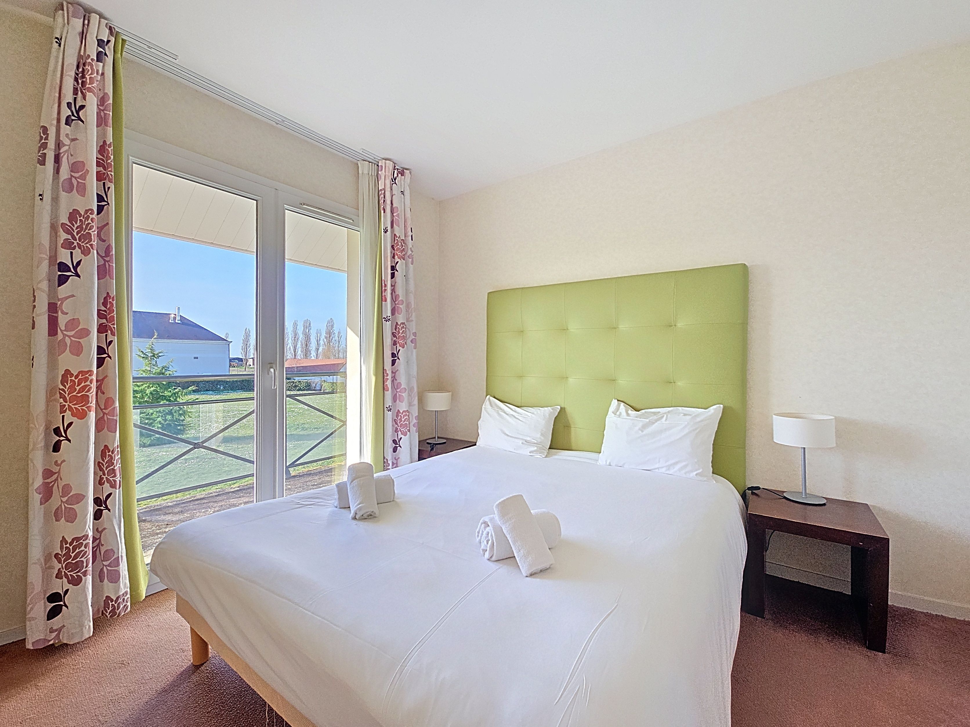 image Upstairs double bedroom with green headboard, white linens, and flowery curtains beside garden-view glass doors