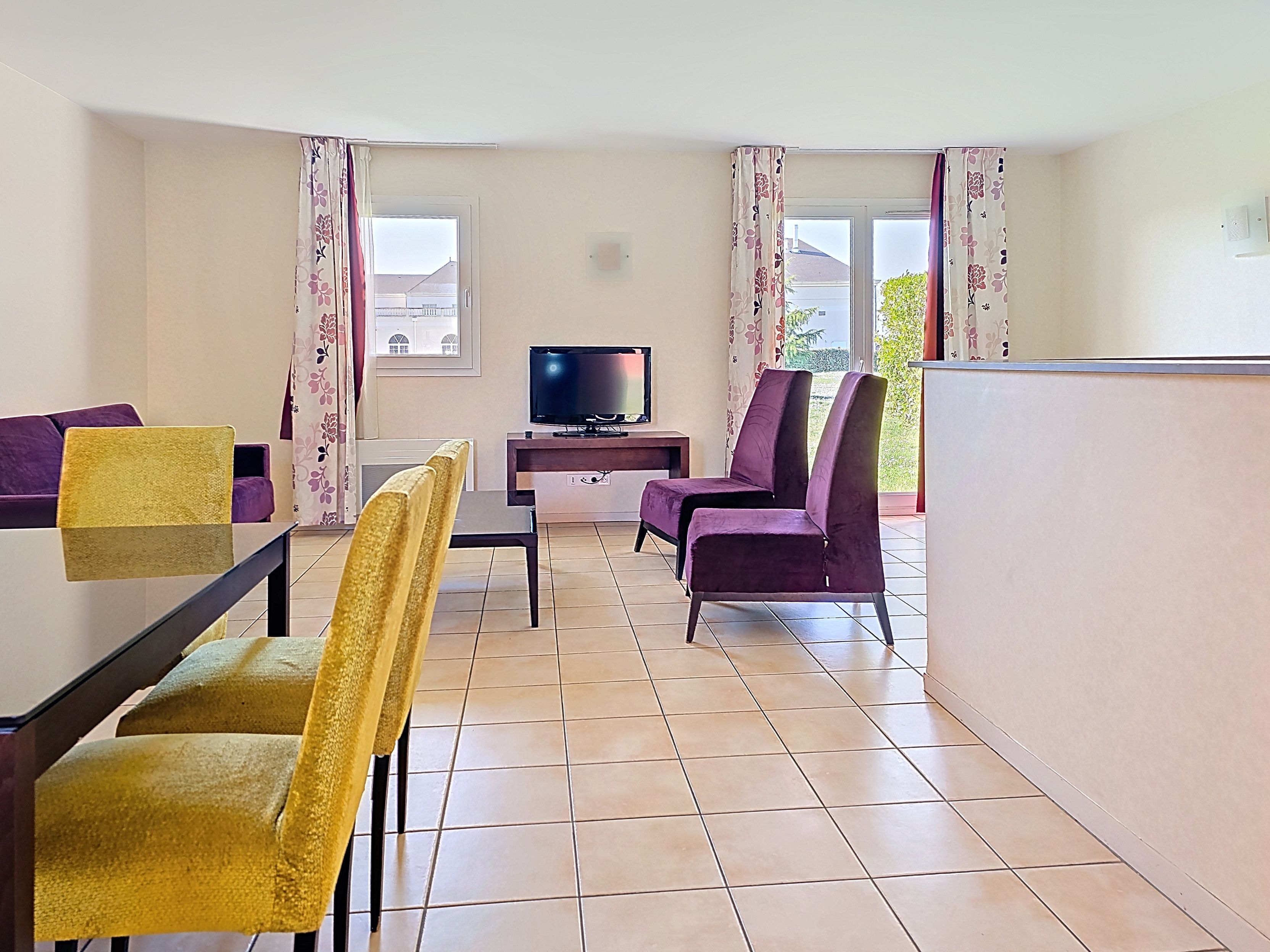 image Open living space with purple sofa, glass dining table, and yellow chairs — natural light from large windows