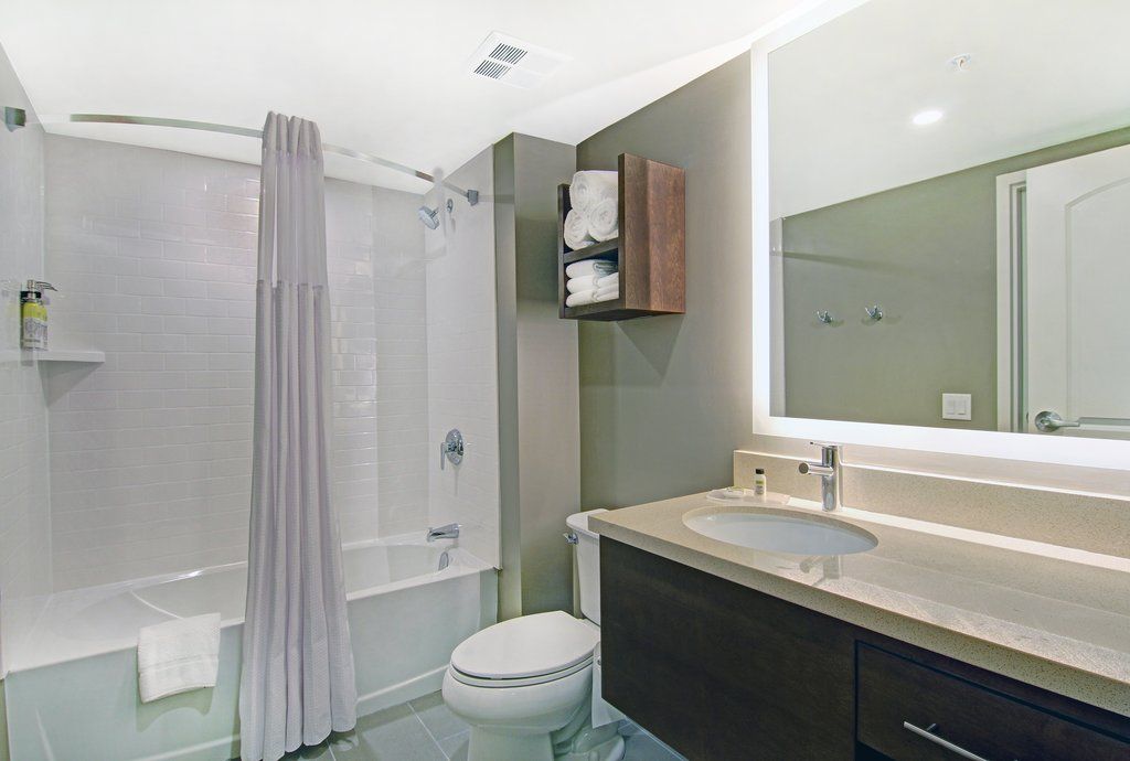 image White-tiled shower with clear curtain, dark wood vanity, integrated sink, and shelves stocked with white towels — accessible bathroom layout.