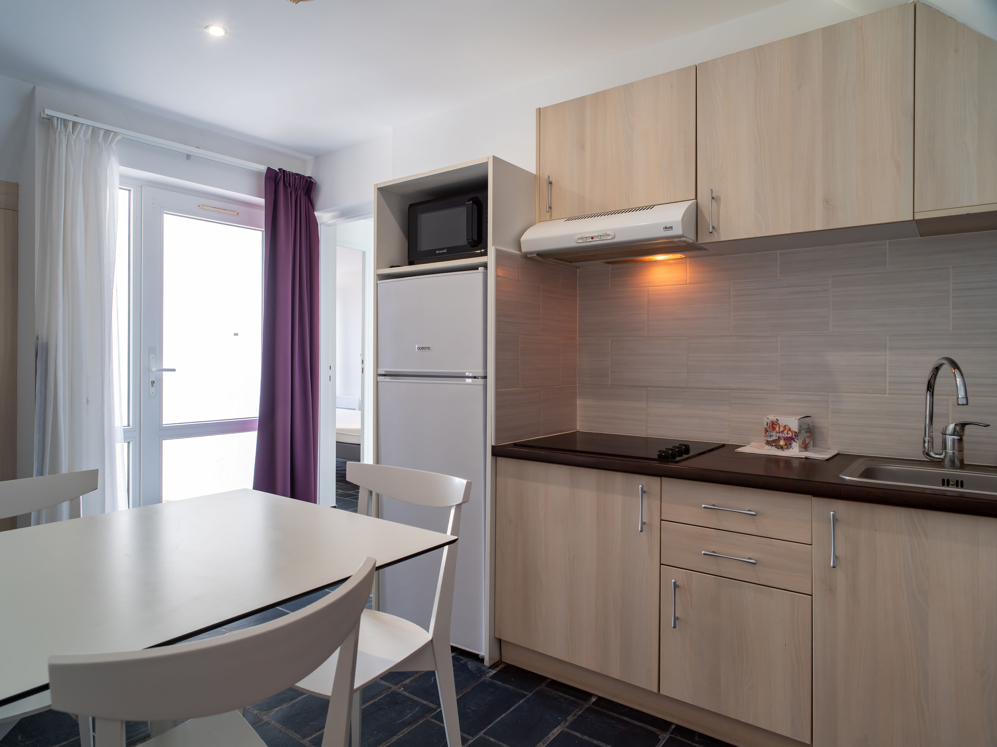 image Light-wood kitchenette with ceramic hob, microwave, fridge-freezer, and dishwasher — round dining table seats the group after a day at the beach.