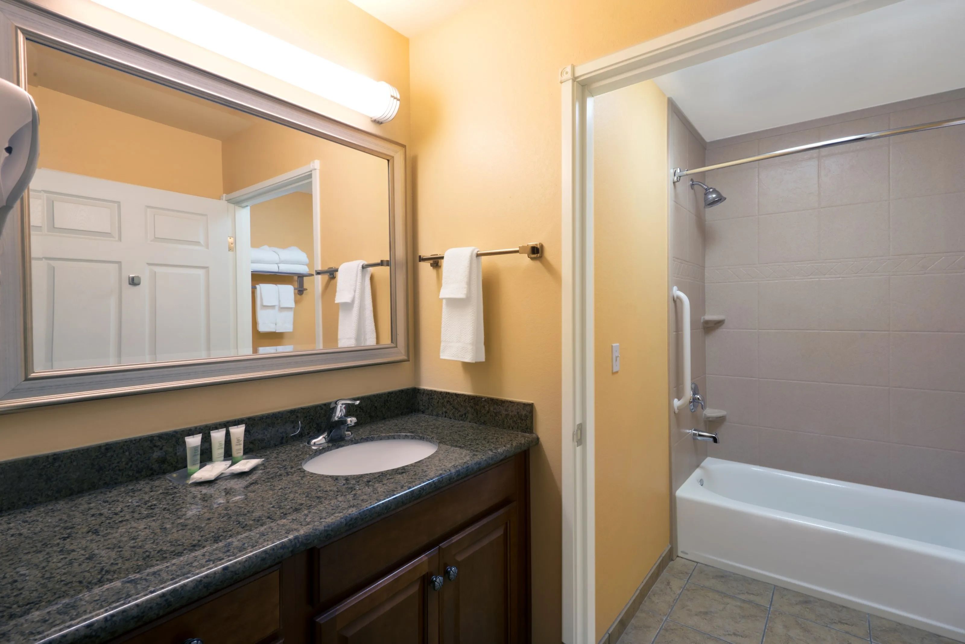 image Bathroom with granite countertop, round sink, toiletries, tub/shower, and white towels. Please note that grab bars may not be present.
