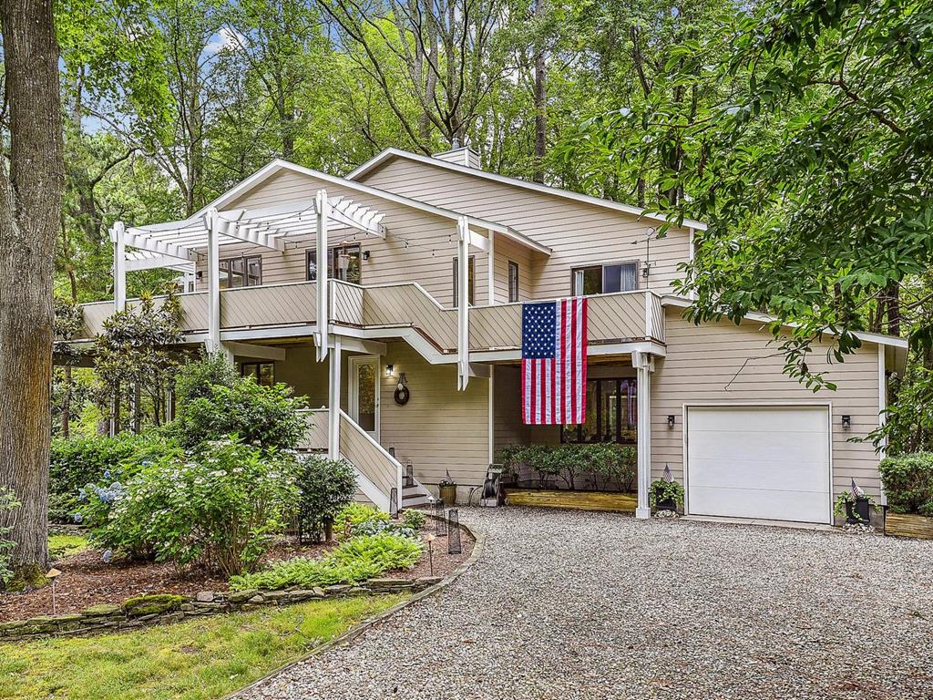 image Full exterior view — gravel driveway, one-car garage, pergola deck above, and American flag at the entrance