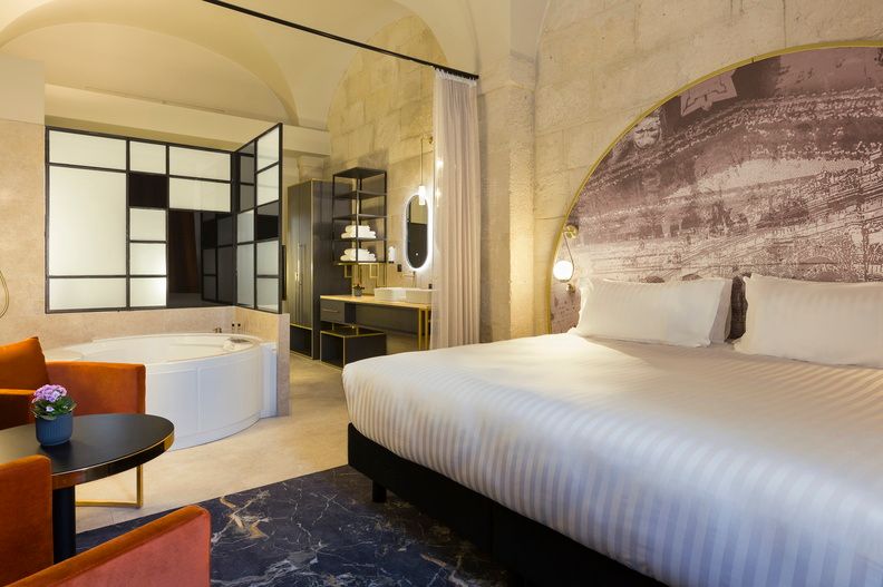 image The deluxe room featuring a king-size bed, a dramatic arched fresco headboard wall, and an open-plan bathroom behind elegant black glass partitions.