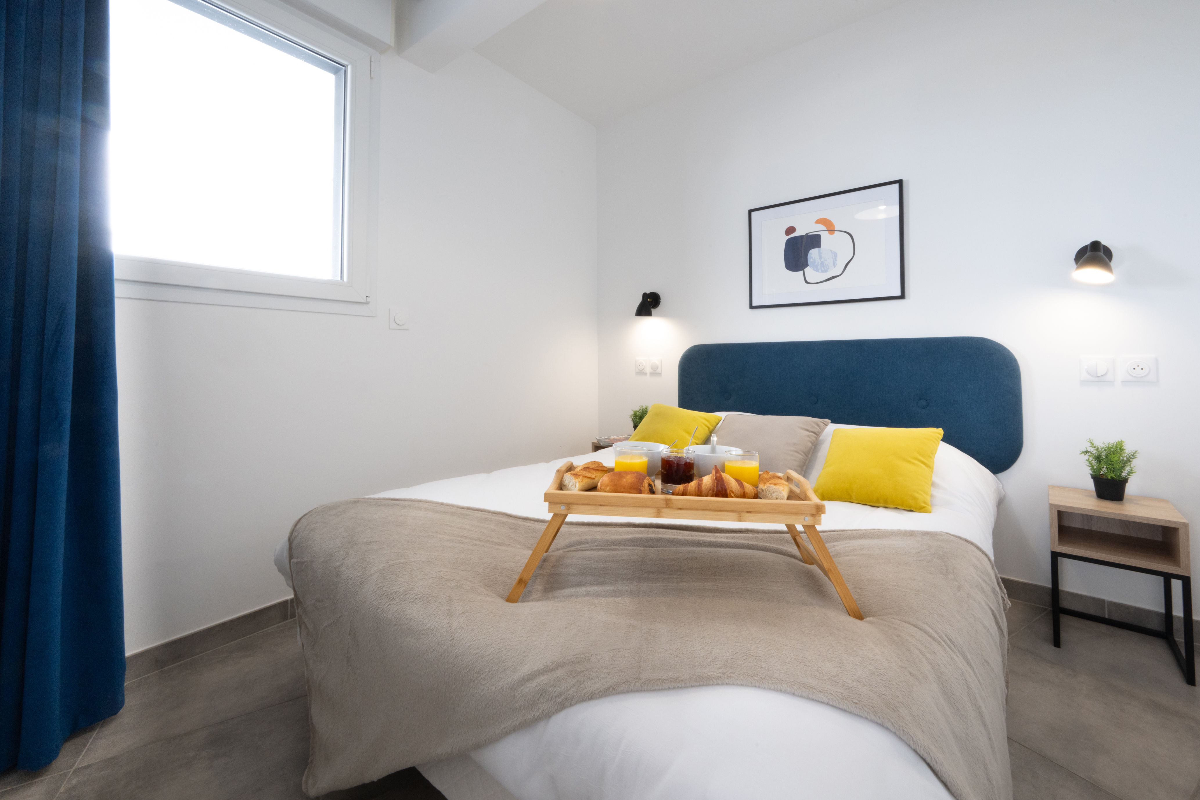 image Double bed with a blue headboard, yellow cushions, and wall sconces — the bedroom opens to natural light through blue curtains.