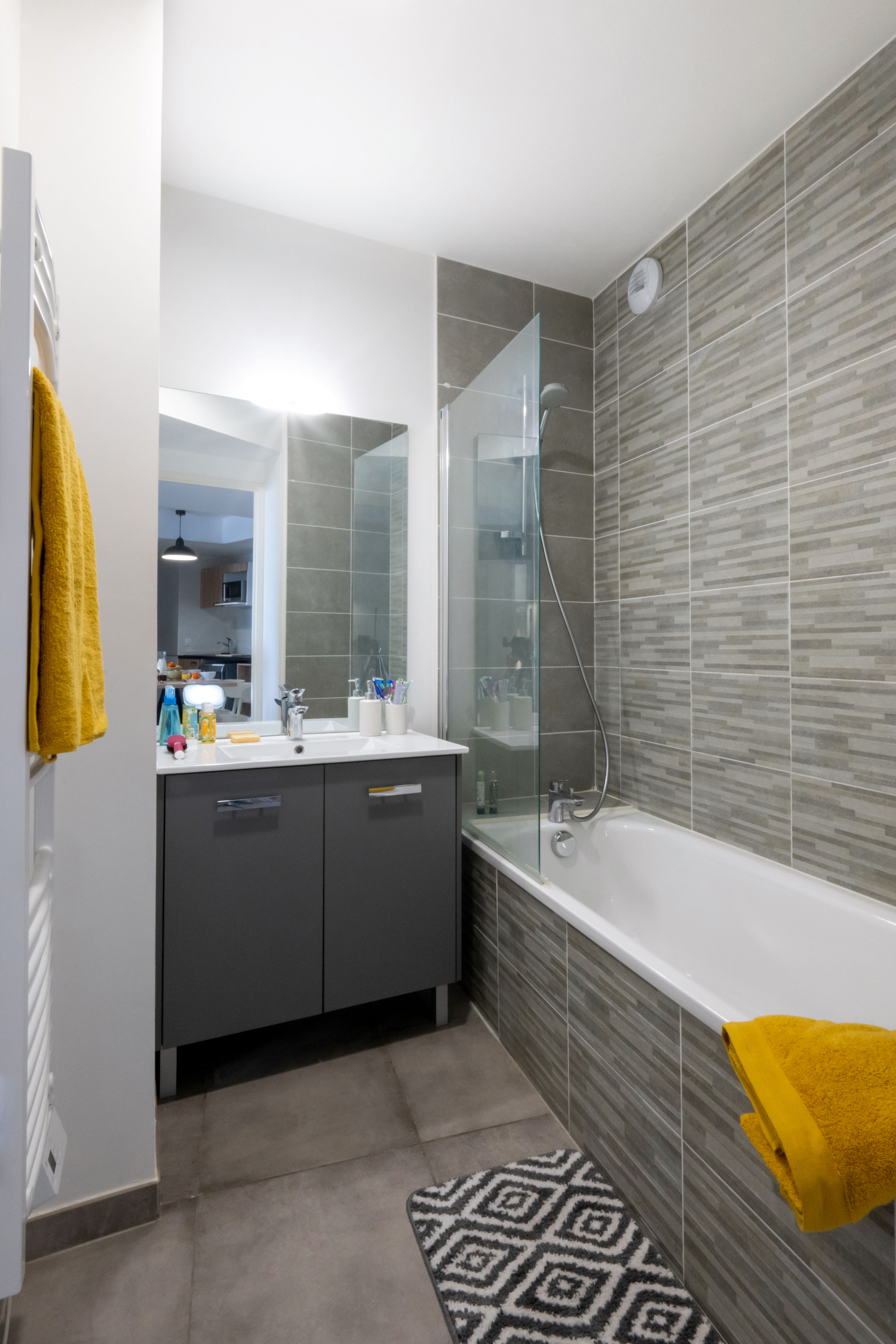 image Gray-tiled bathroom with a bathtub, glass shower screen, and yellow towel detail against a dual-tone tile surround.