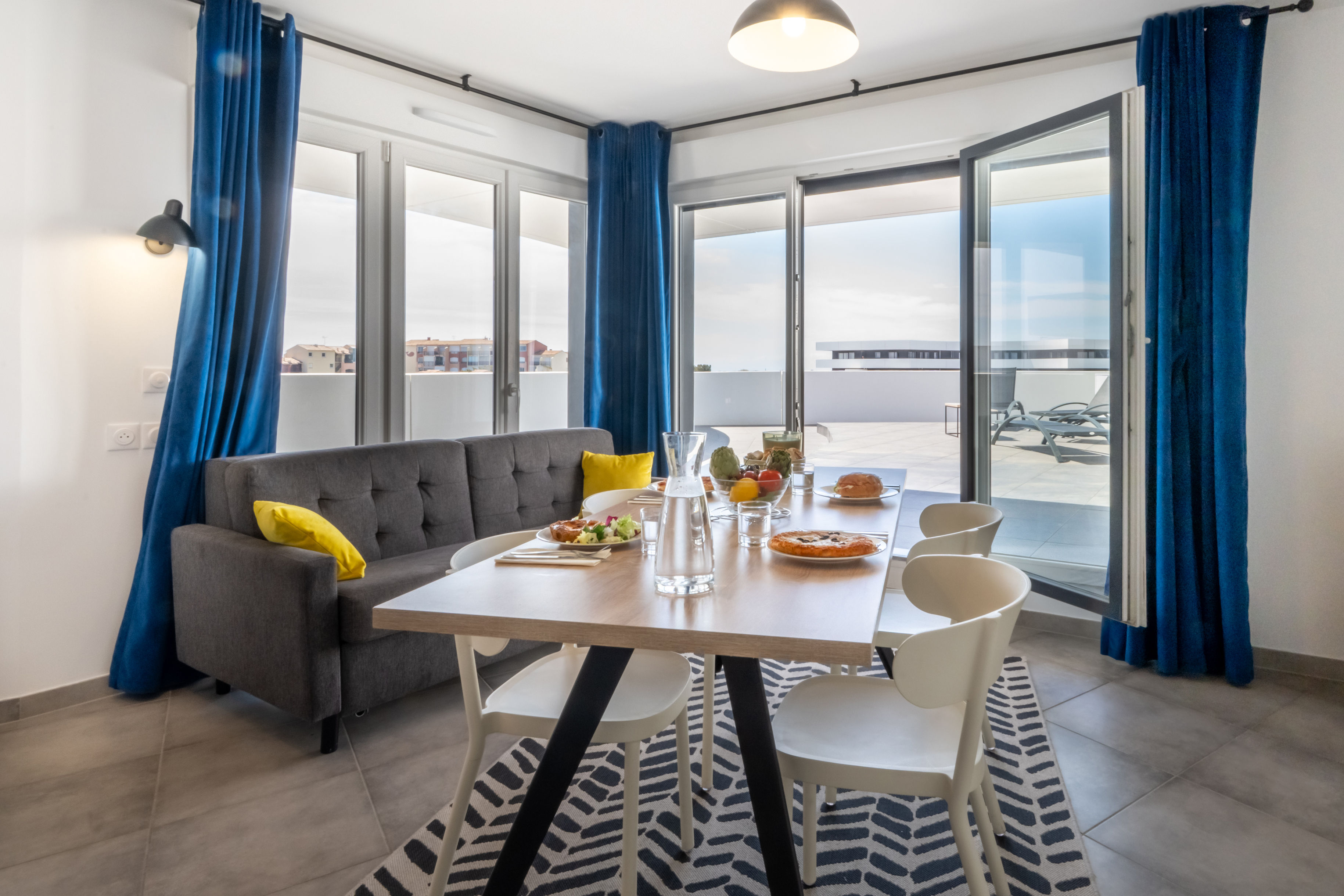 image Open-plan living and dining area with wooden table, gray sofa, yellow cushions, and blue curtains — space to spread out after a day at the marina.