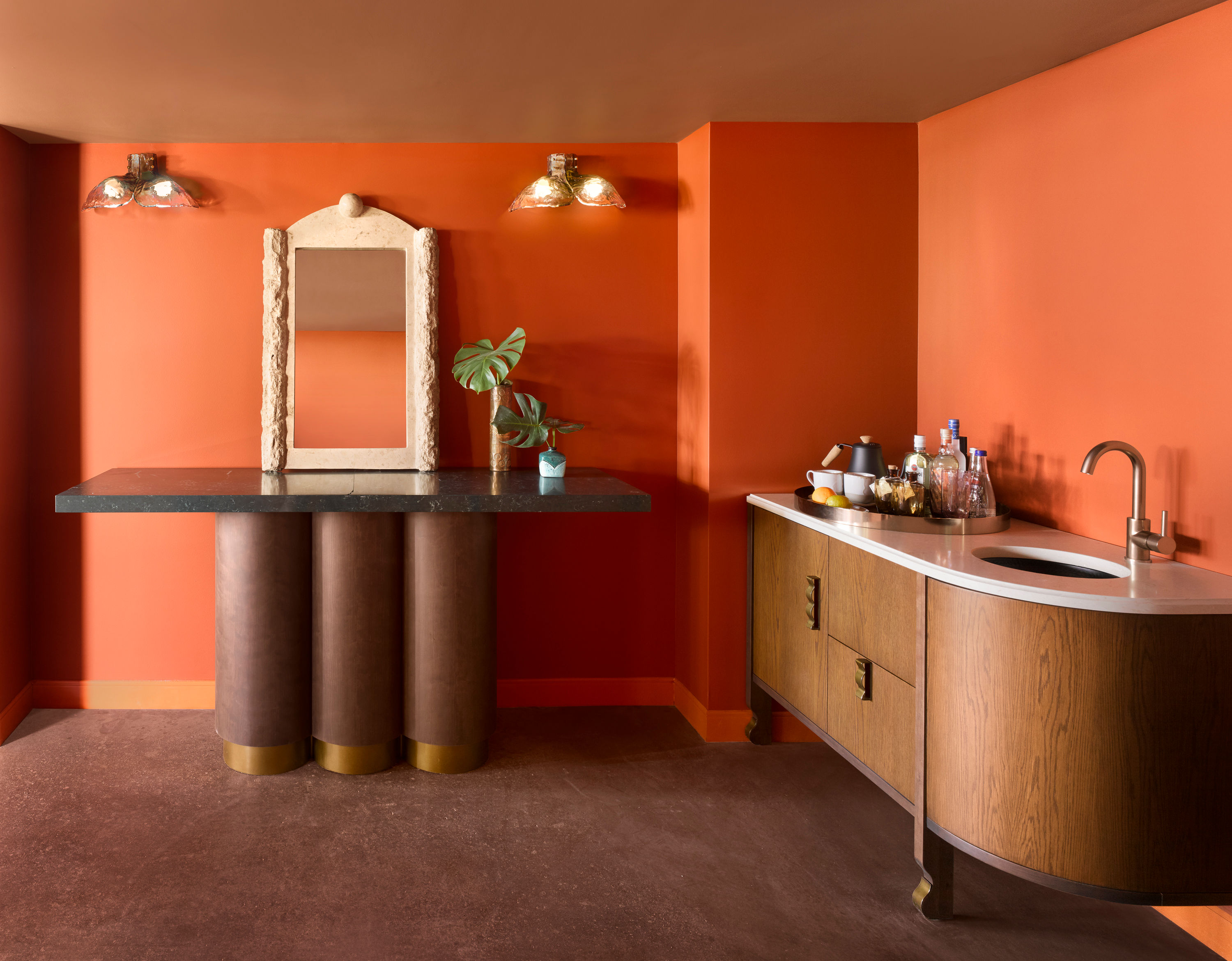 image A stone-topped console table with cylindrical bases stands against a bold orange wall with a large round mirror