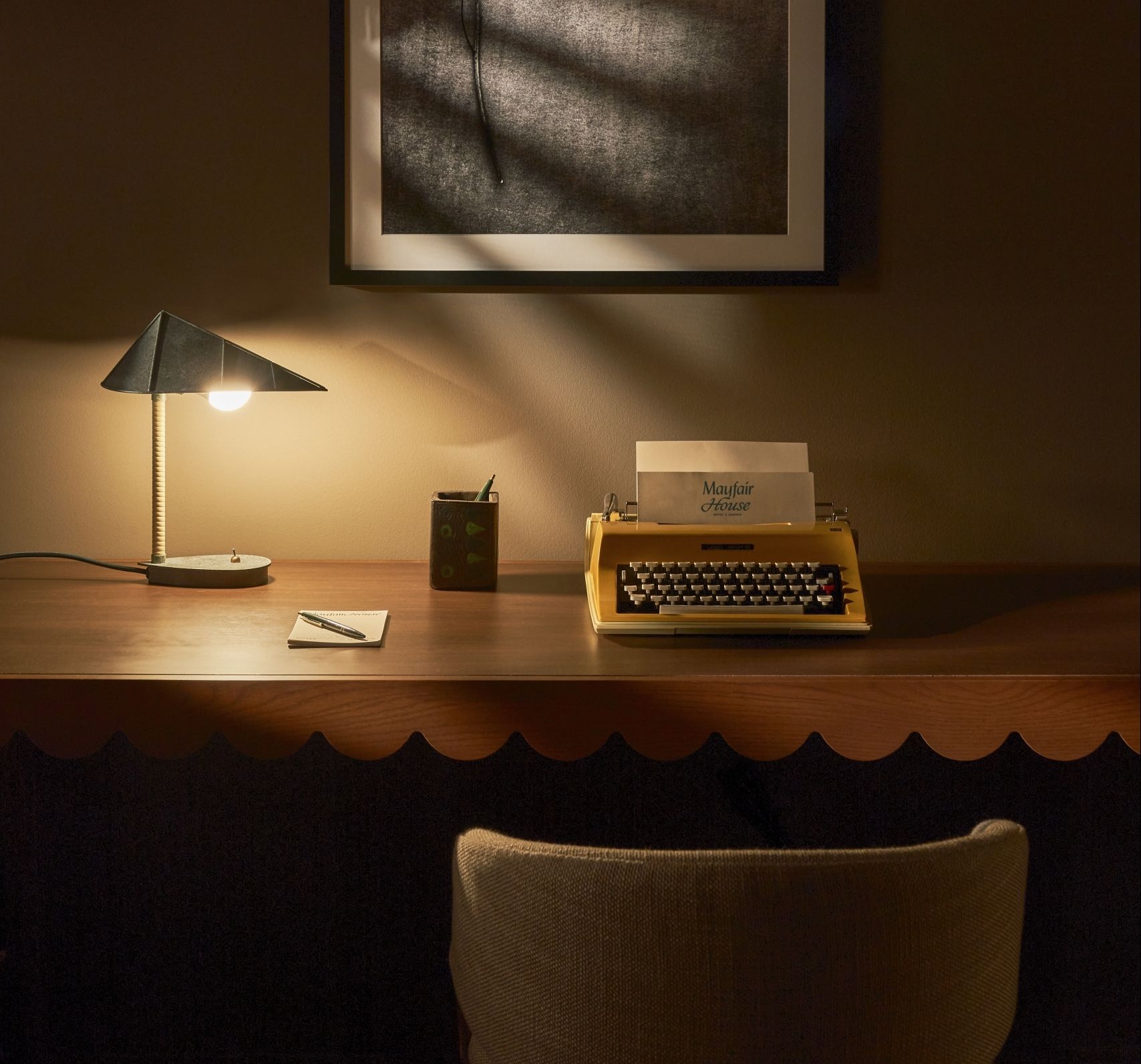 image Scallop-edged walnut desk with a yellow typewriter, open notepad, and a round-bulb lamp in a dim corner