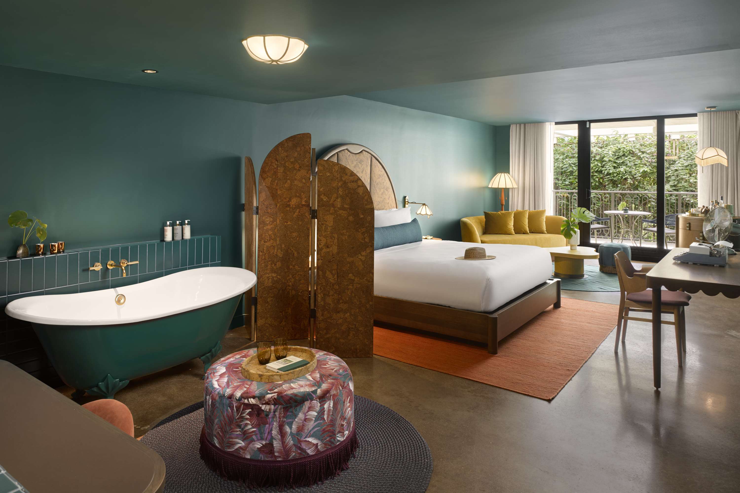 image King bed with white linens beside a white freestanding tub on a teal base, with a yellow sofa and tropical pouf in the room
